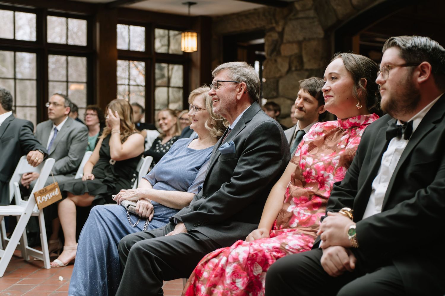 Wedding guests seated in rustic venue, smiling and watching ceremony, dressed in formal attire including floral dress and tuxedo.