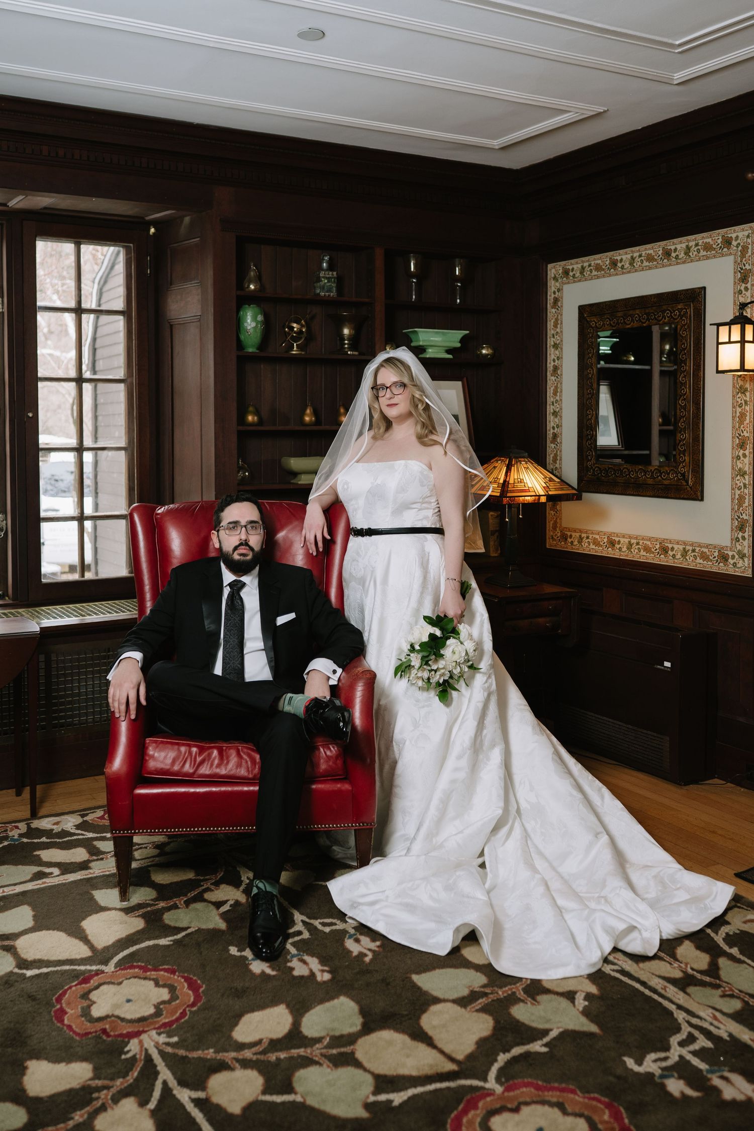 Bride in white gown with veil stands beside groom in black suit seated in red leather chair in elegant wood-paneled room.