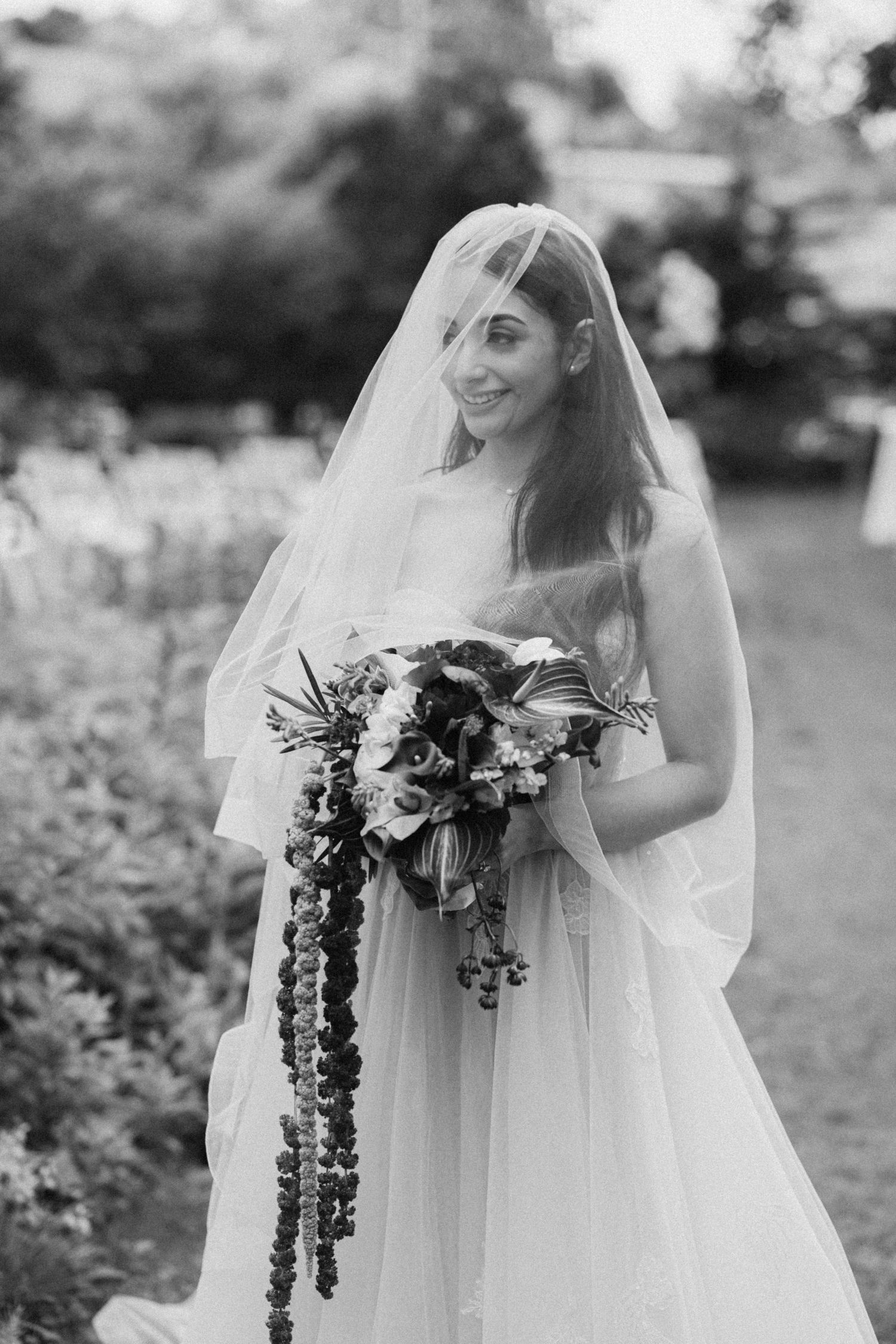 Black and white bridal portrait of a smiling bride in a veil holding a cascading floral bouquet outdoors.