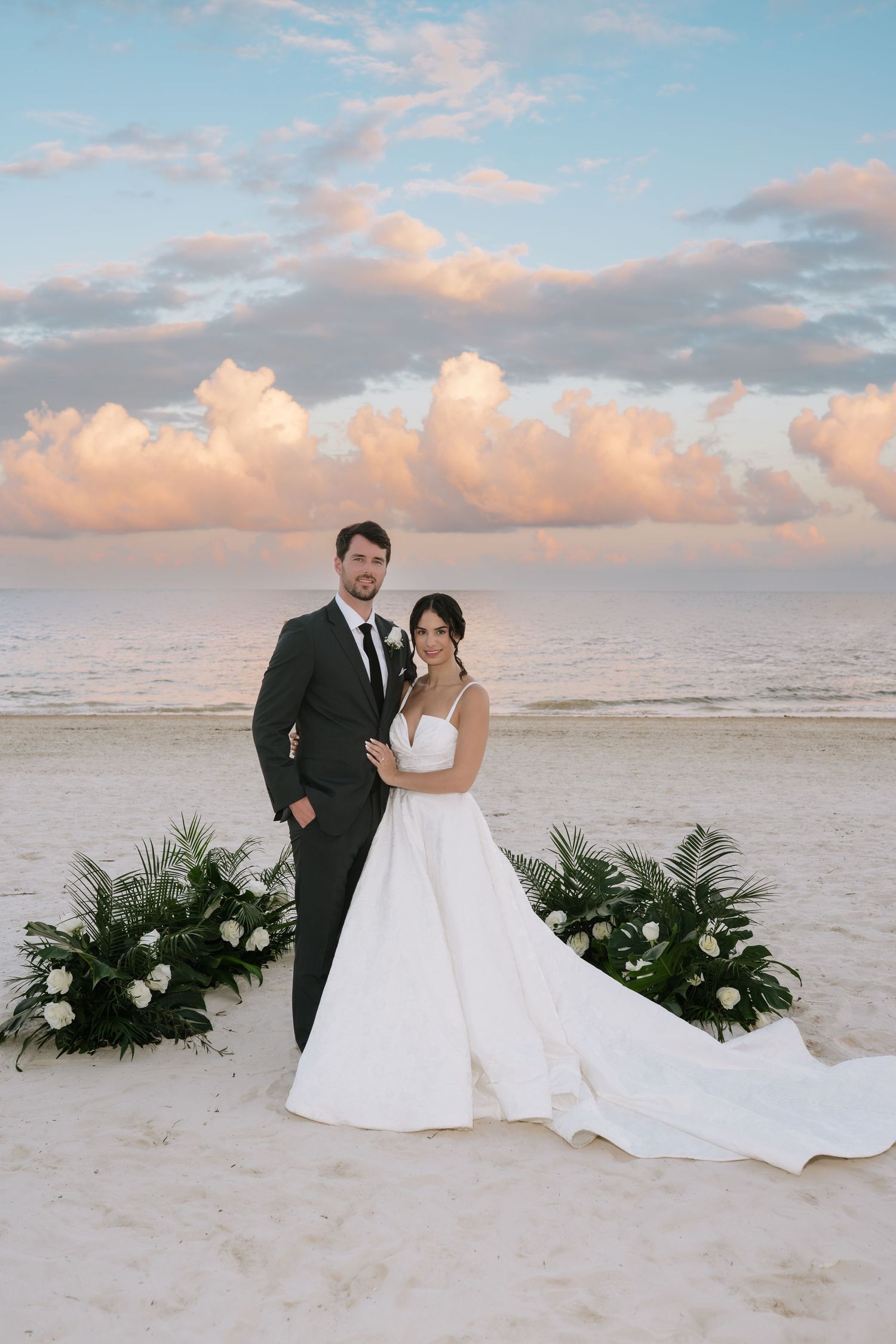 A couple poses for romantic beach wedding photos at sunset with tropical greenery arrangements in the sand.