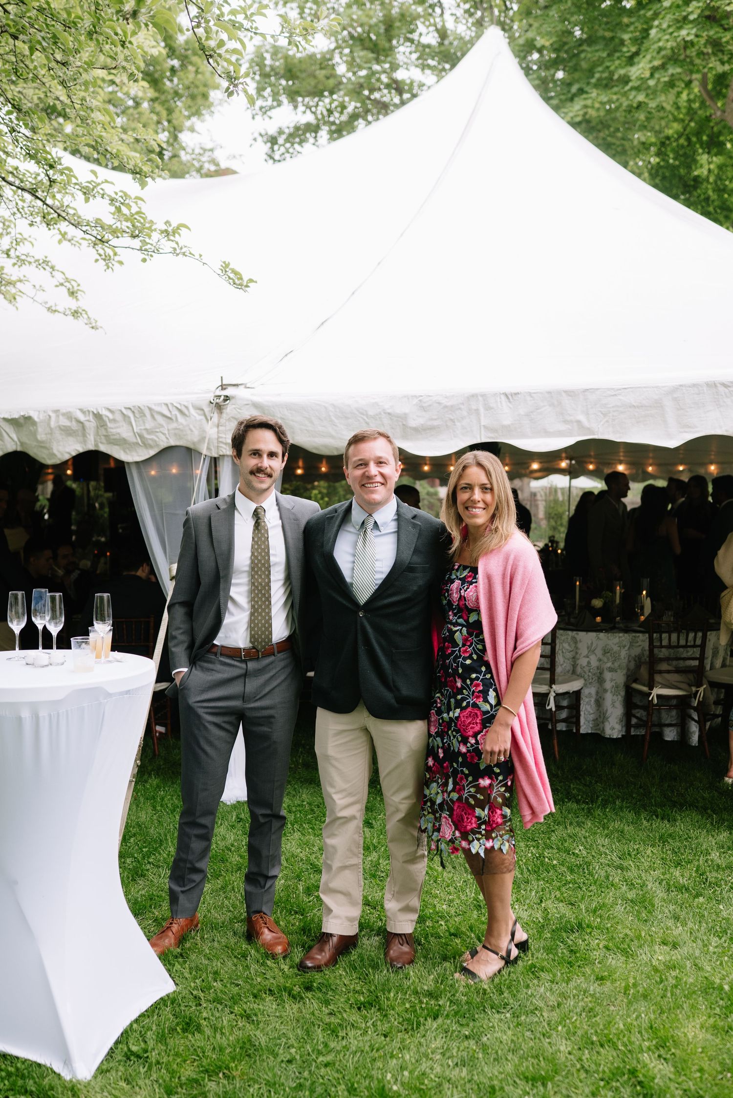 Three guests smile together outdoors at an elegant tented wedding reception on a green lawn.