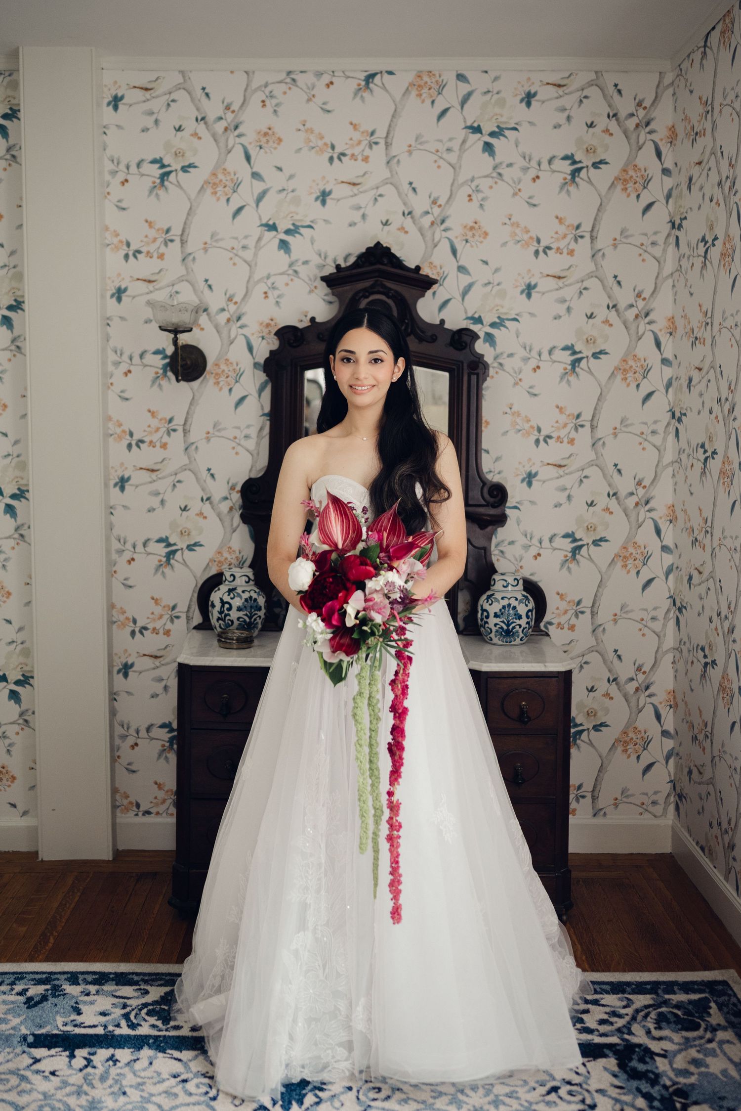 Bride in strapless white ball gown holding cascading red and white bouquet, standing before floral wallpaper.