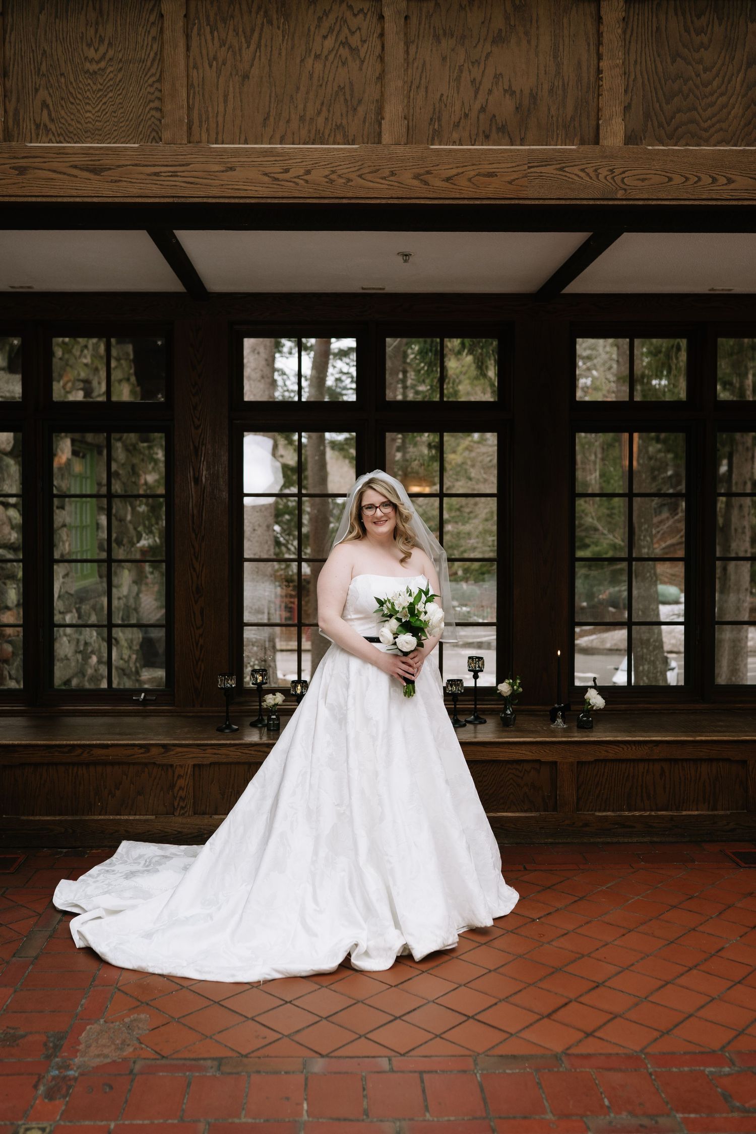 Bride in white ballgown with train holds bouquet indoors before large windows on brick floor.