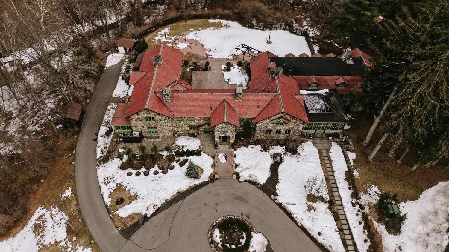 Aerial winter view of a grand stone mansion with red roof surrounded by snow-covered trees and circular driveway.