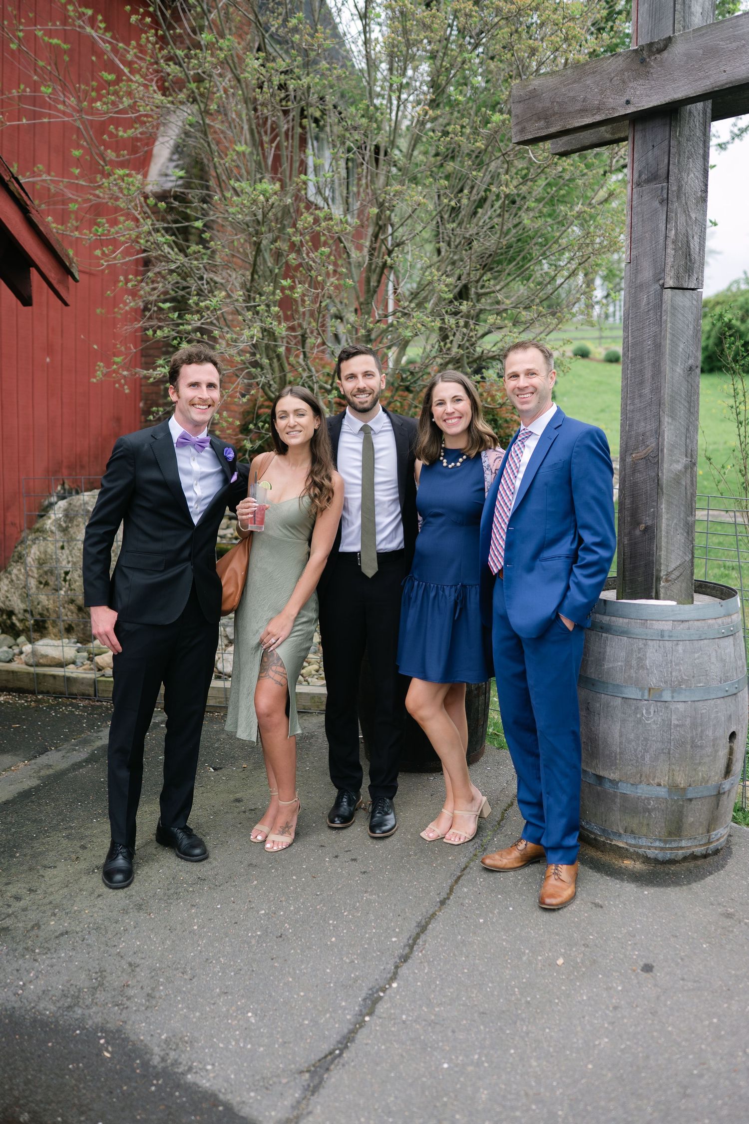 Wedding party poses for photos outside a rustic red barn with wooden barrels.