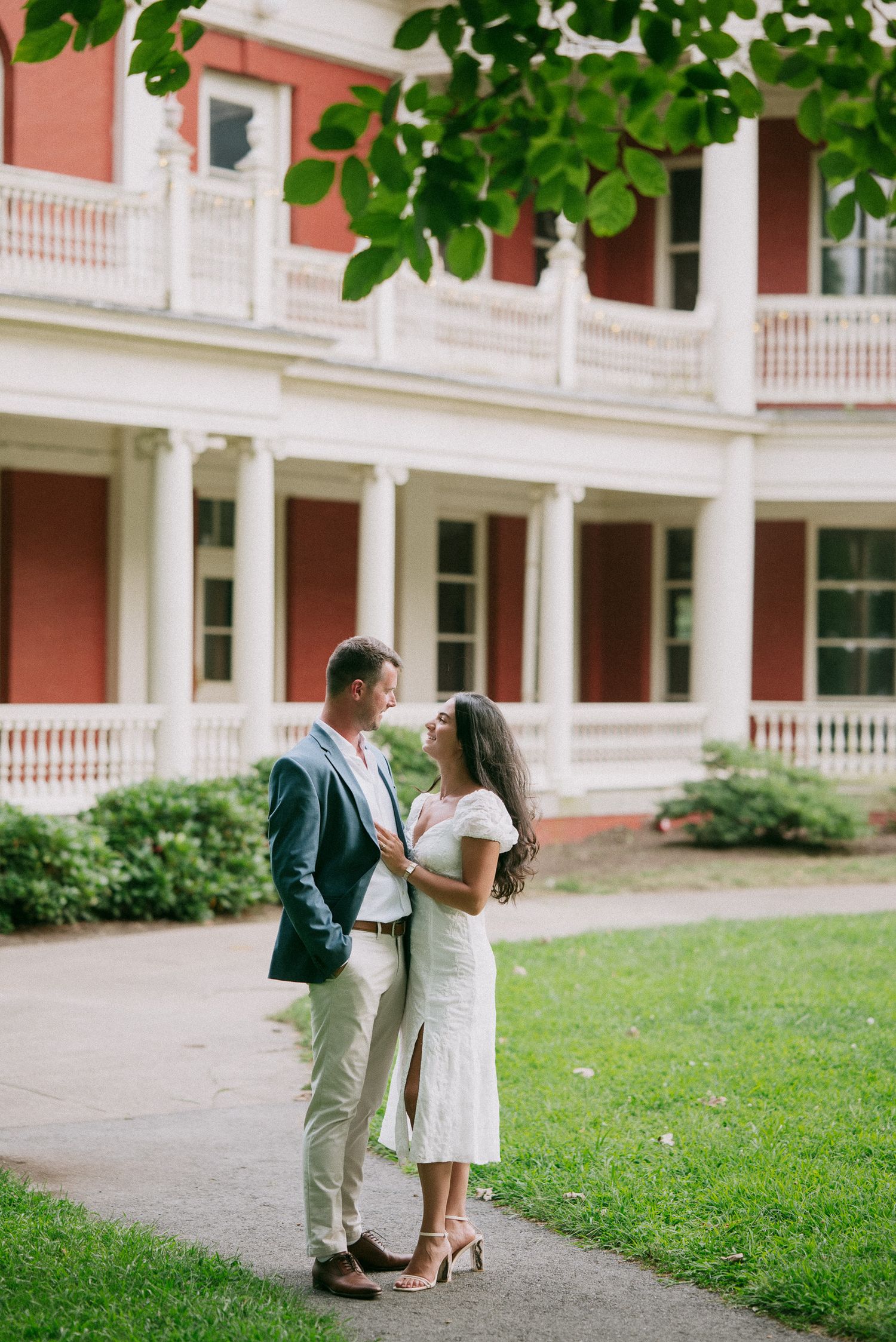 Couple in formal attire posing together on a lawn with a grand colonial style building in the background.