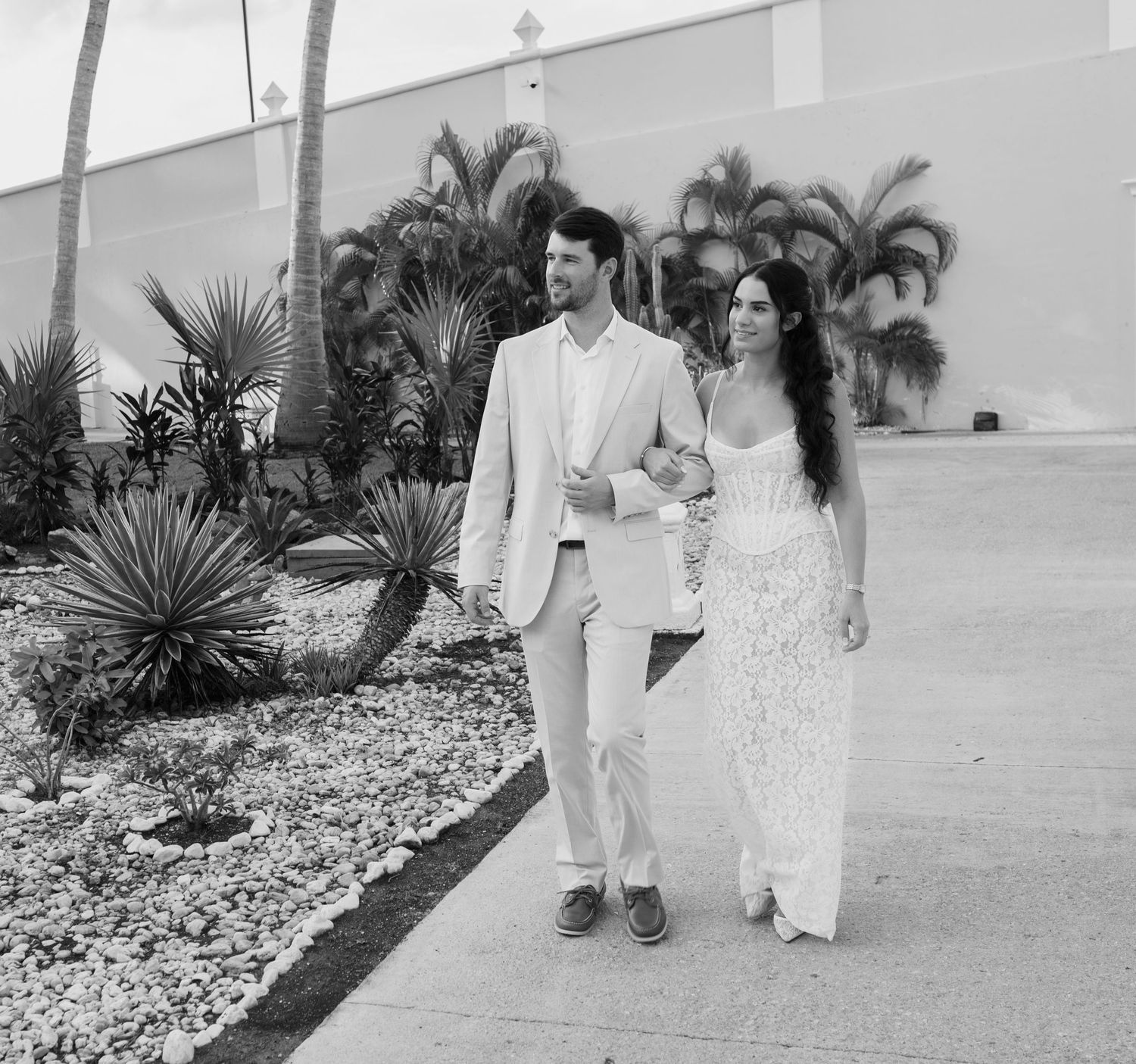 A couple in wedding attire walks together on a path lined with tropical palm trees and landscaping.