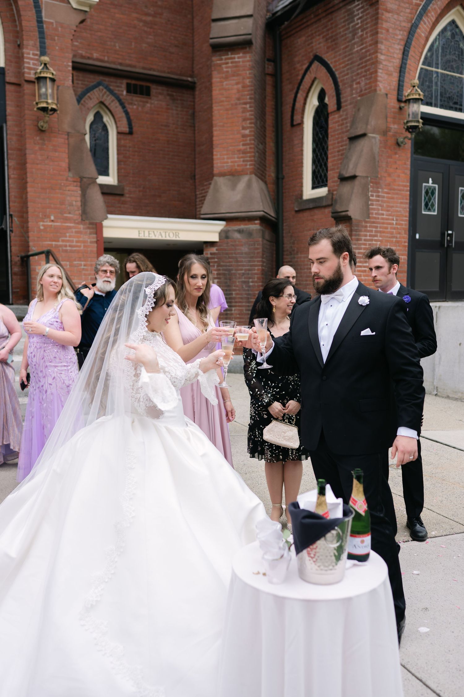 Wedding celebration outside a brick church as attendees gather around a table for a champagne toast.