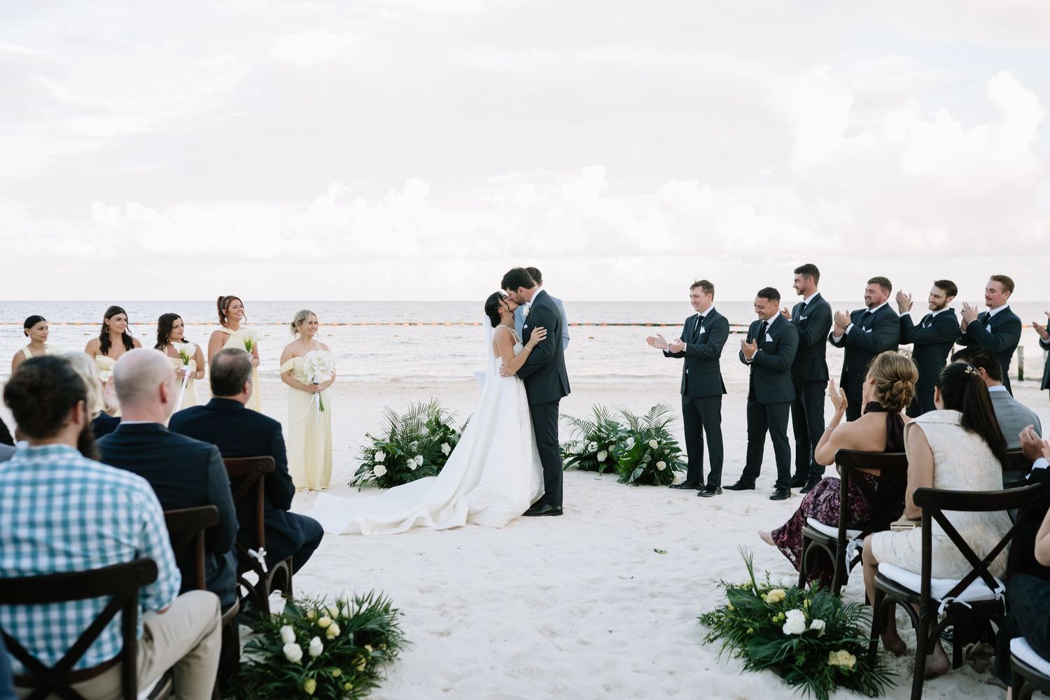 Wide angle view of an outdoor beach wedding ceremony with seated guests and tropical landscaping.