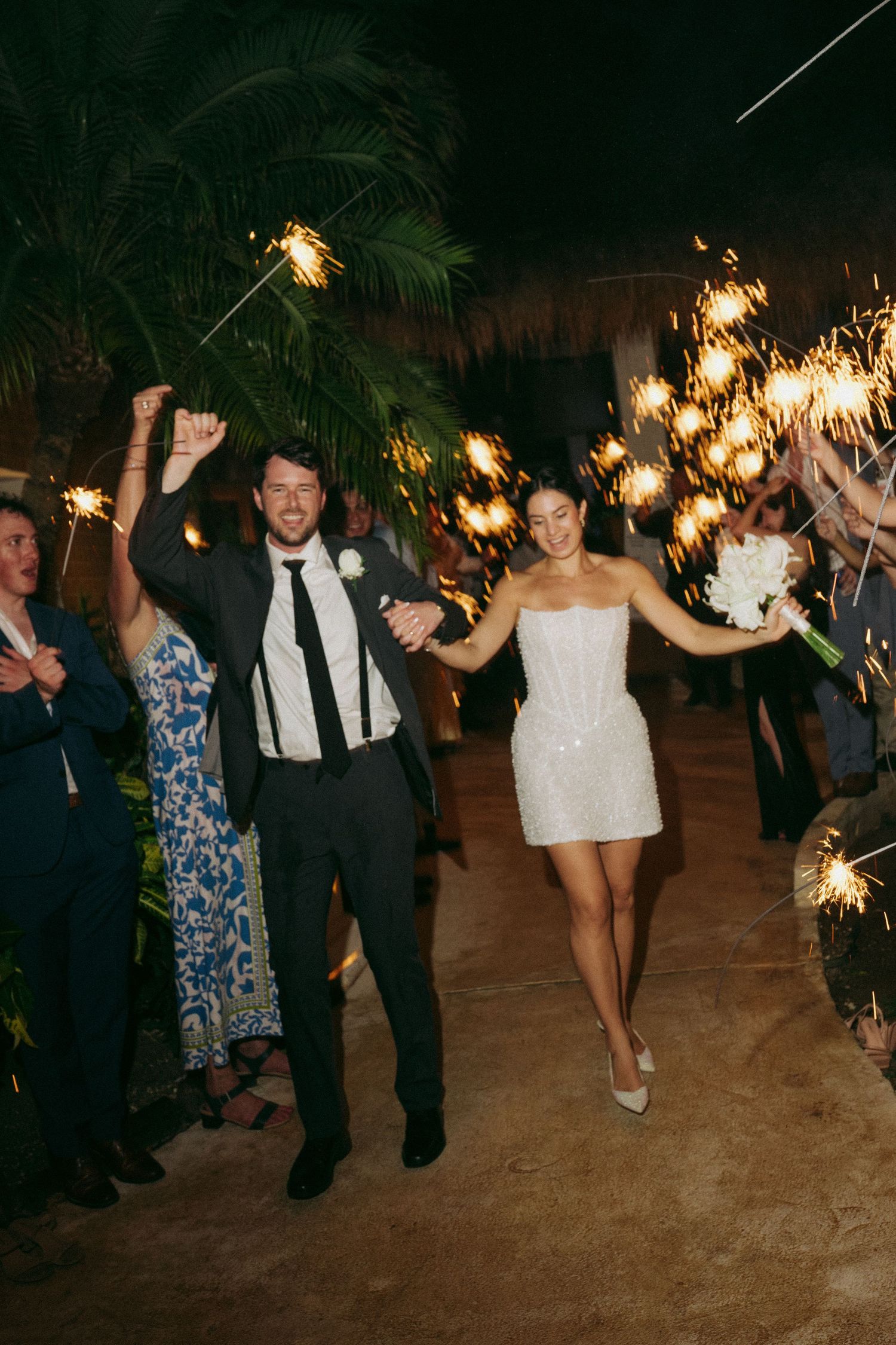 Wedding guests holding sparklers while dancing outdoors at night under decorative string lights.