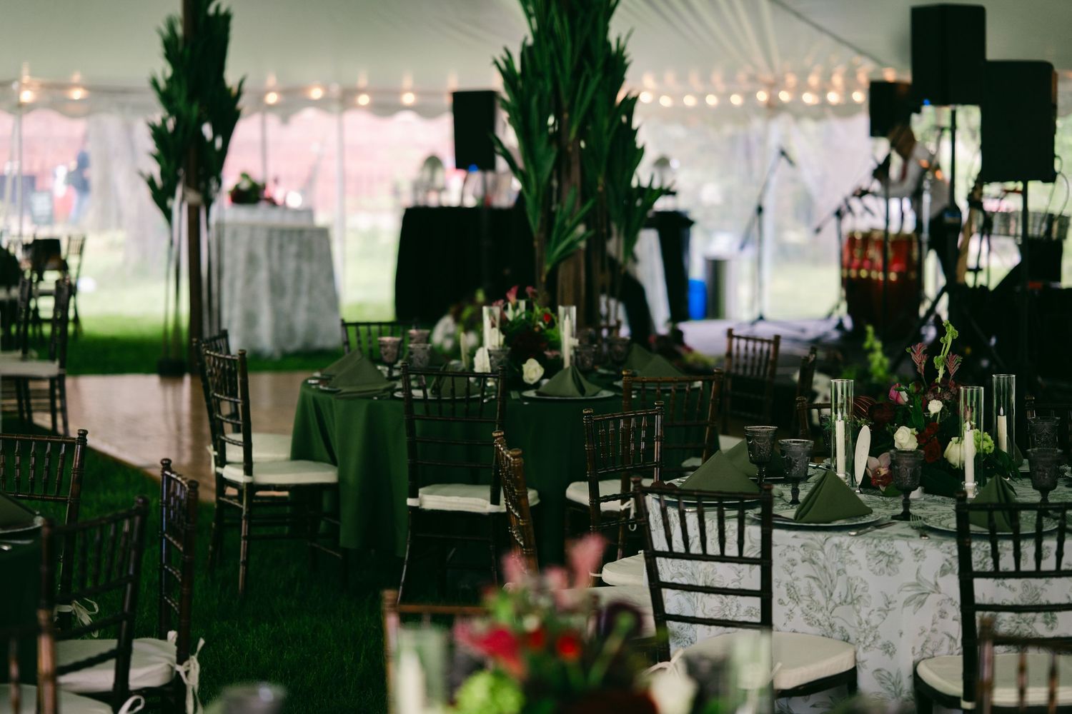 Elegant wedding reception tent with dark chiavari chairs, green tablecloths, tall floral arrangements, and string lights overhead.