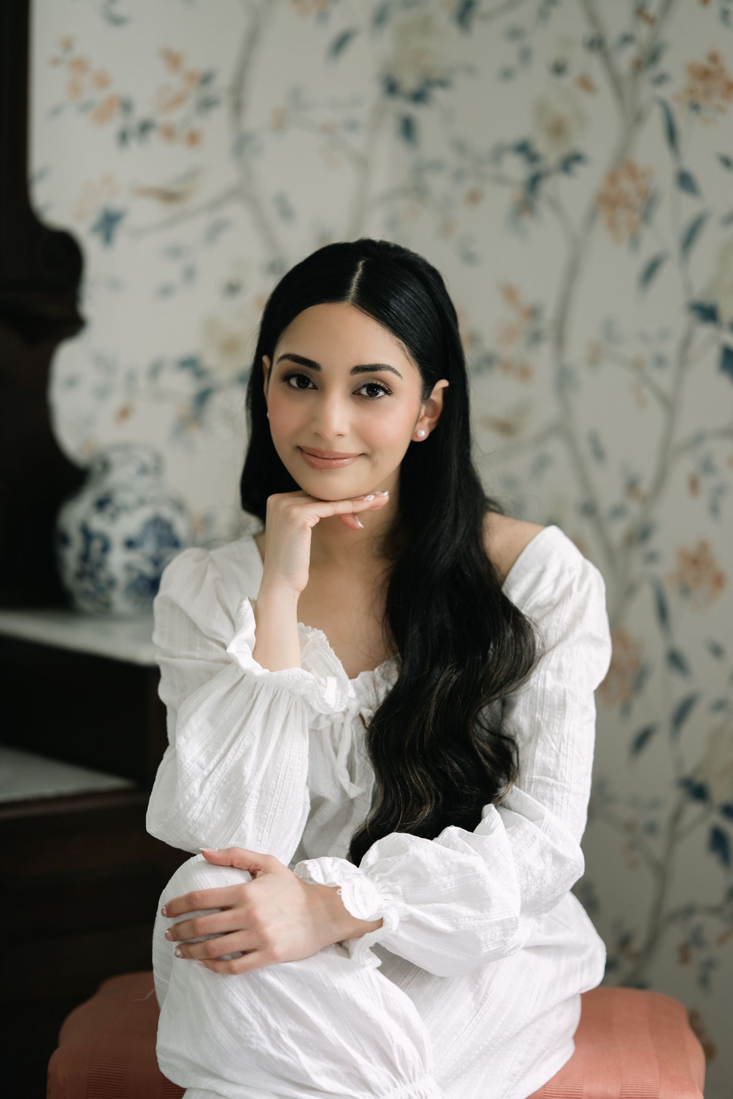 Young woman in white off-shoulder dress with long dark hair poses gracefully against floral wallpaper background.
