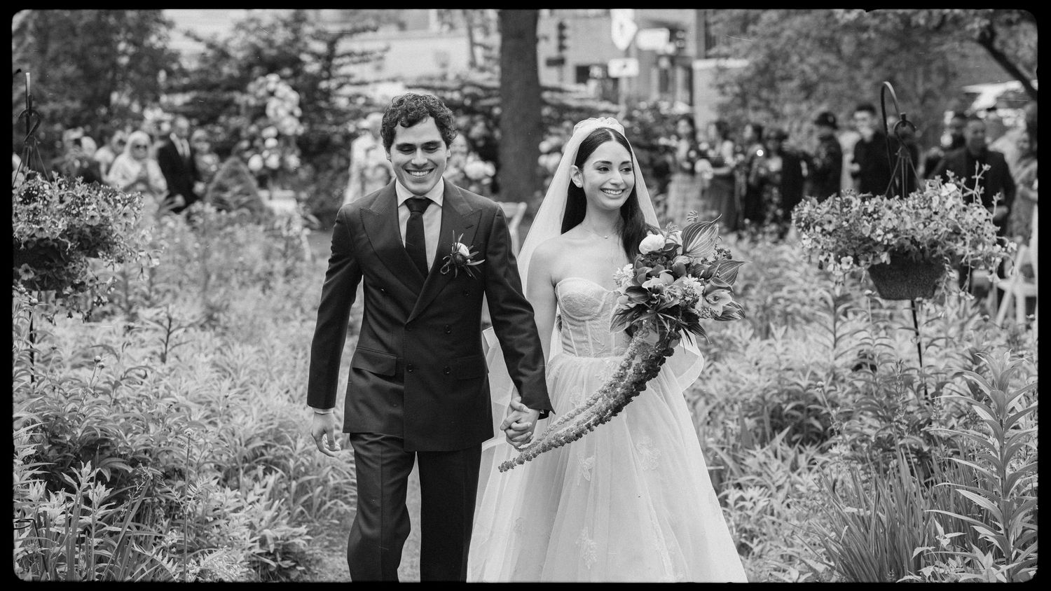 Black and white photo of a smiling couple holding hands walking through a garden on their wedding day.
