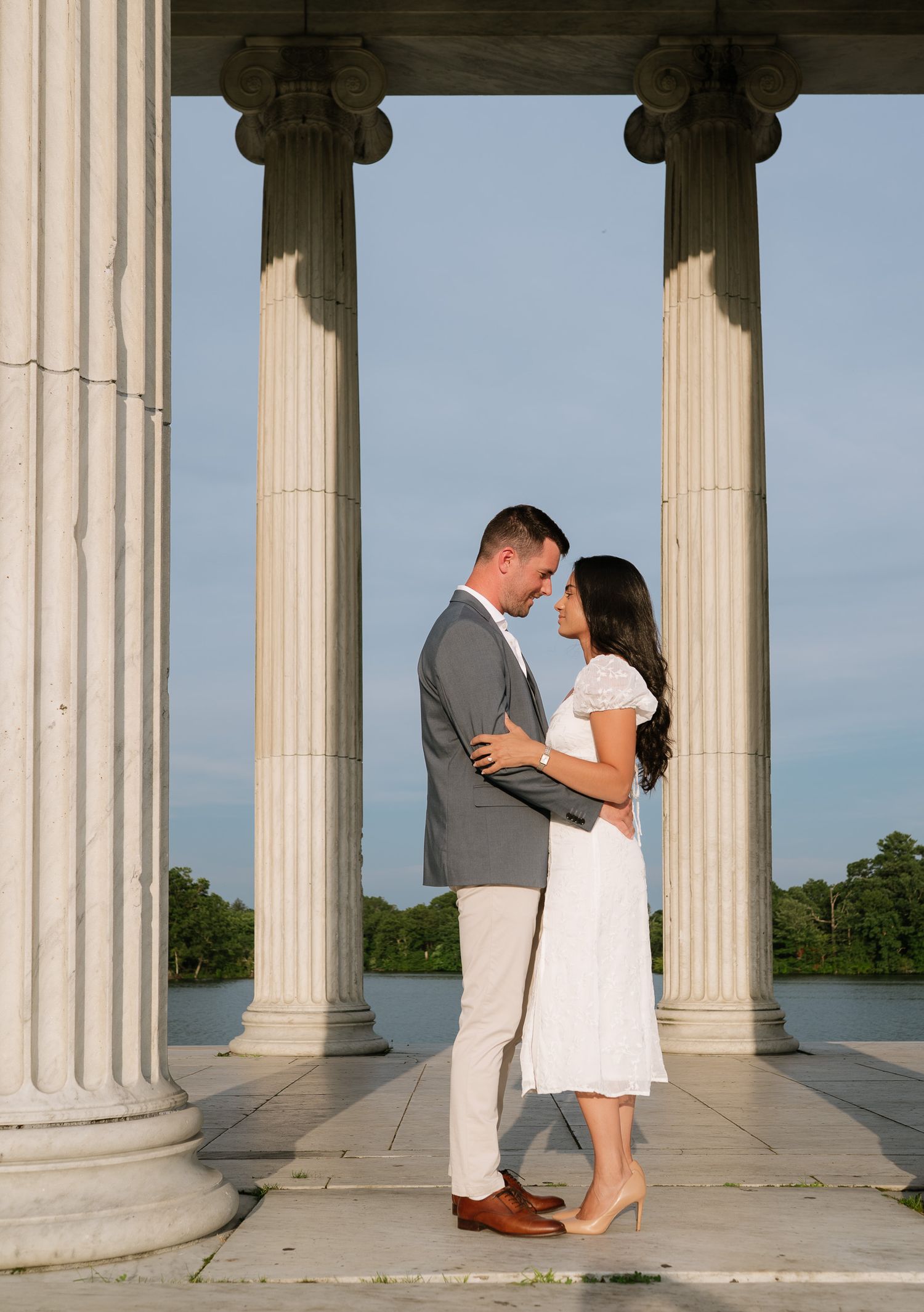 A couple embraces between elegant white columns of a neoclassical building.
