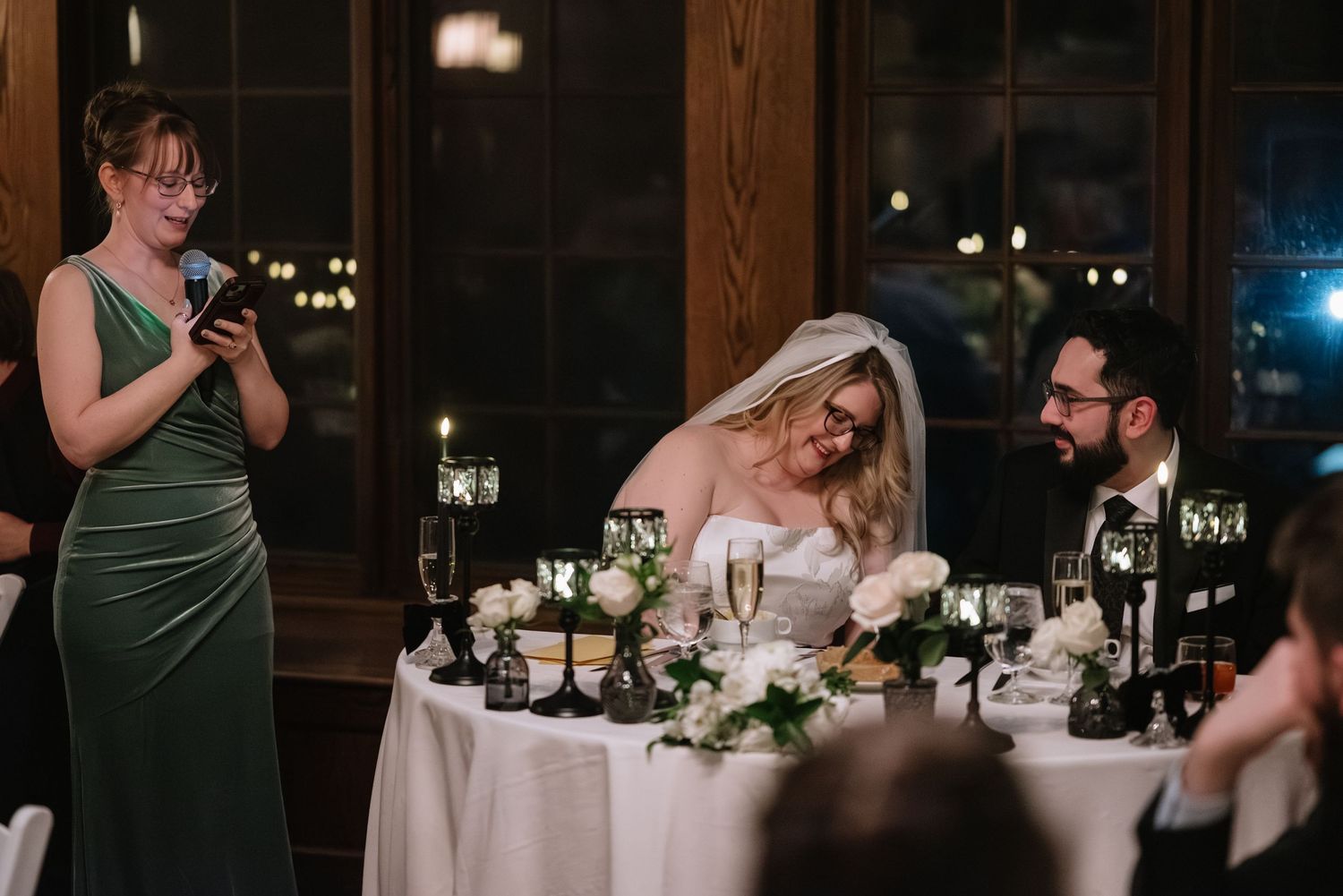Bridesmaid in green gown gives a heartfelt speech as bride and groom smile at wedding reception table.