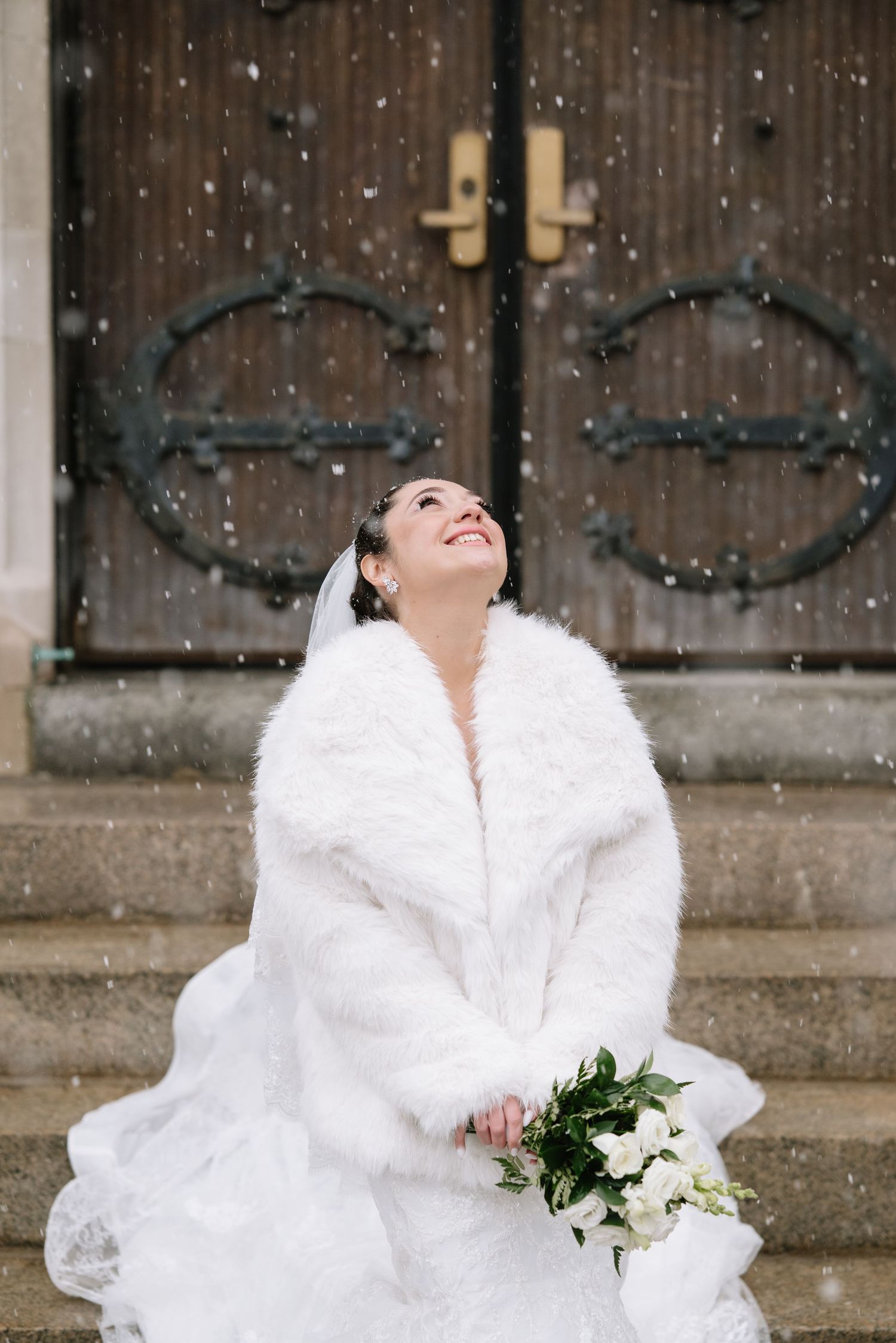 Joyful winter bride in white fur wrap laughs in the falling snow while holding a white floral bouquet.