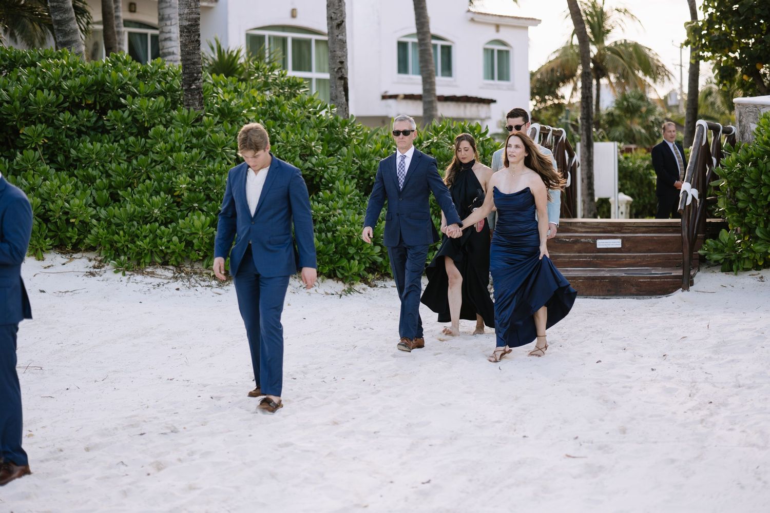 Wedding party members walk along a white sandy beach path with palm trees and buildings in the background.