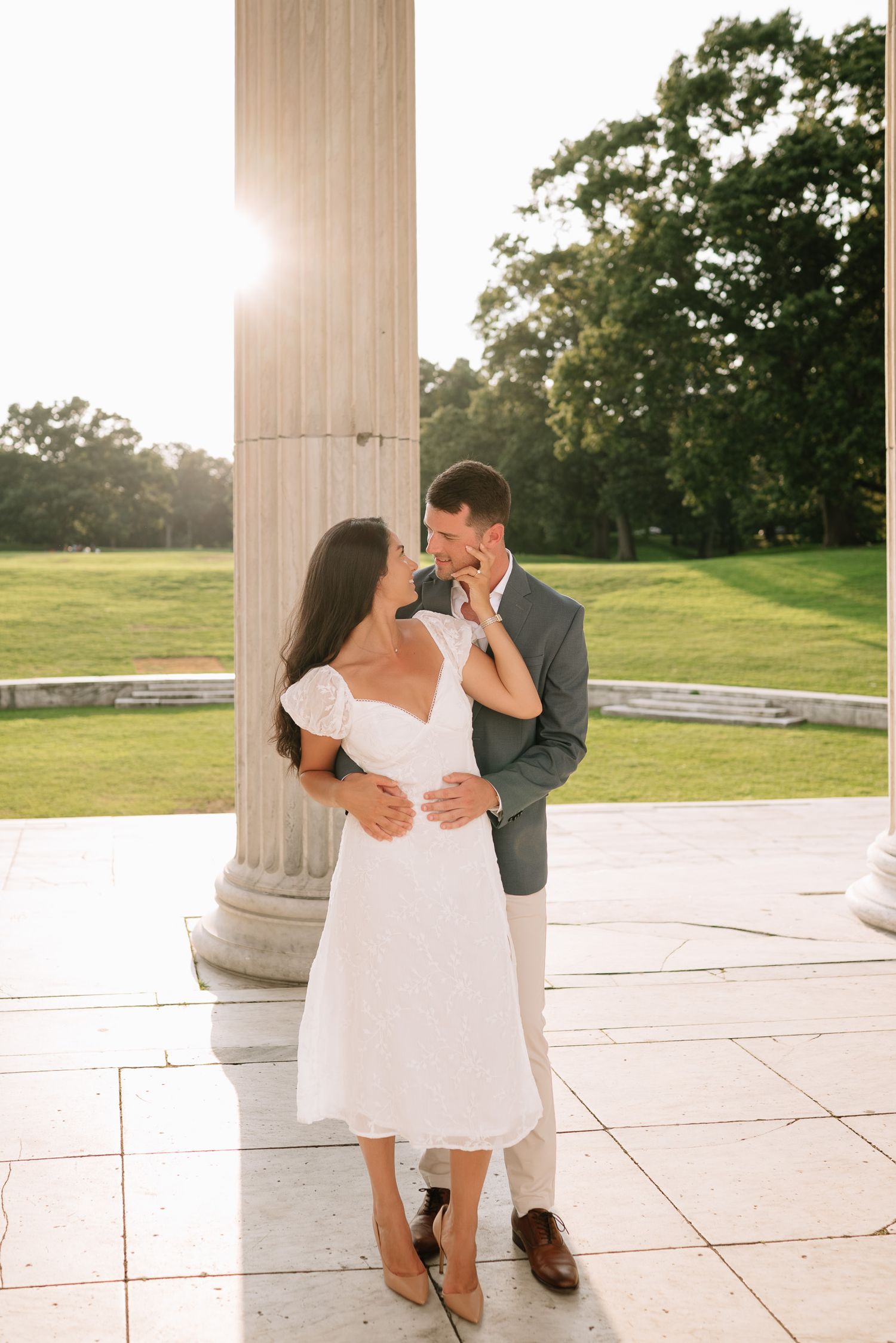A romantic couple in formal attire share intimate moments near a classical stone column at sunset.