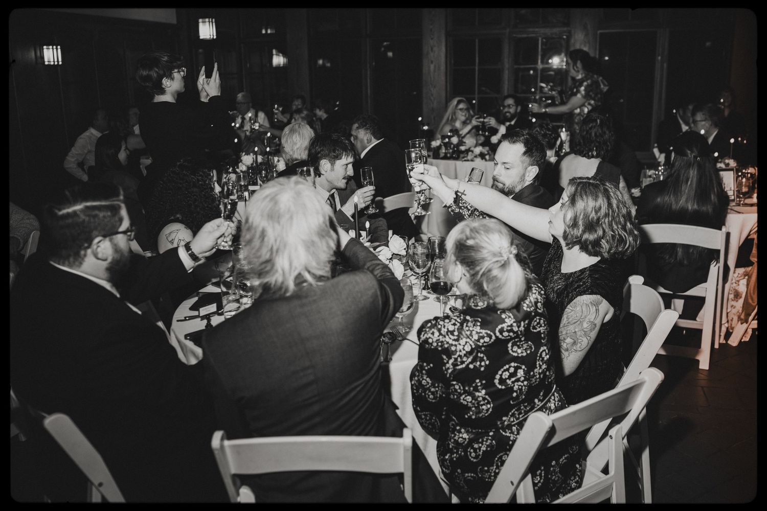 Black and white photo of elegantly dressed guests toasting at a formal dinner event with candles and white chairs.