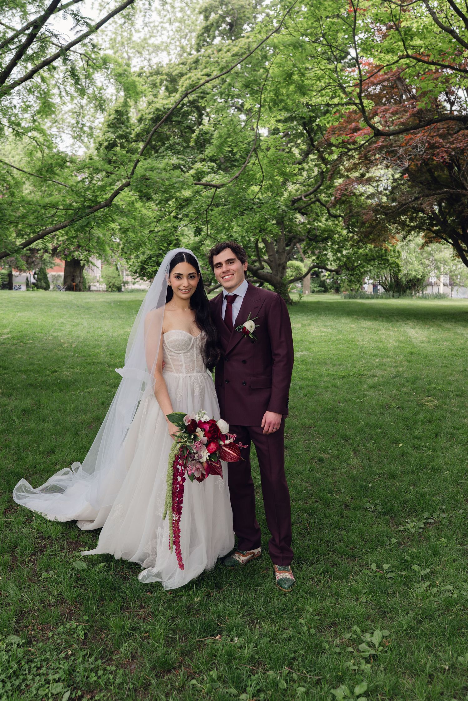 Bride in white gown with veil and groom in burgundy suit pose together outdoors on lush green lawn.