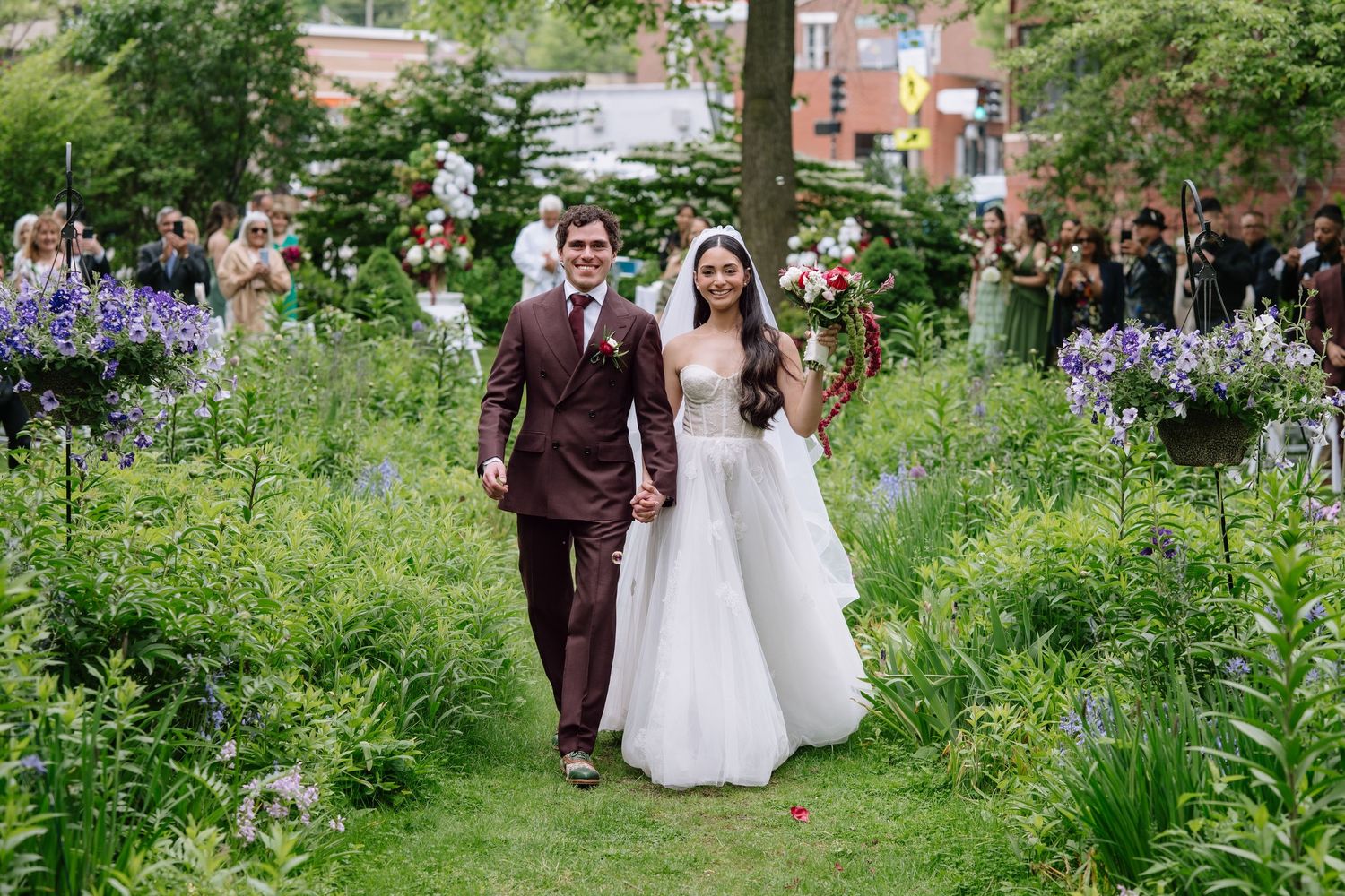 Bride and groom walk through lush garden aisle with purple wildflowers during outdoor wedding ceremony.