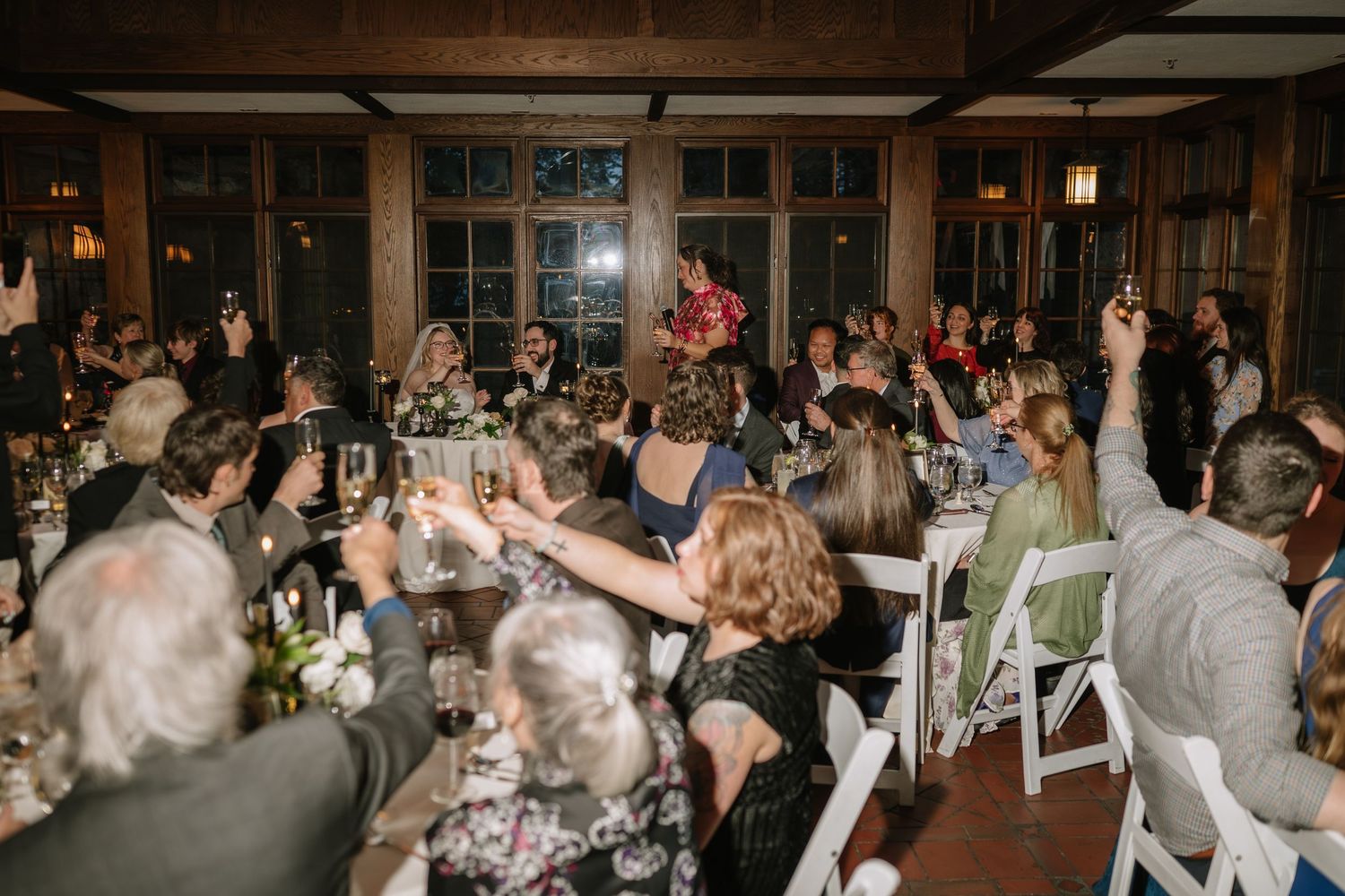 Wedding guests raising glasses in a toast at an elegant indoor reception venue with warm lighting.