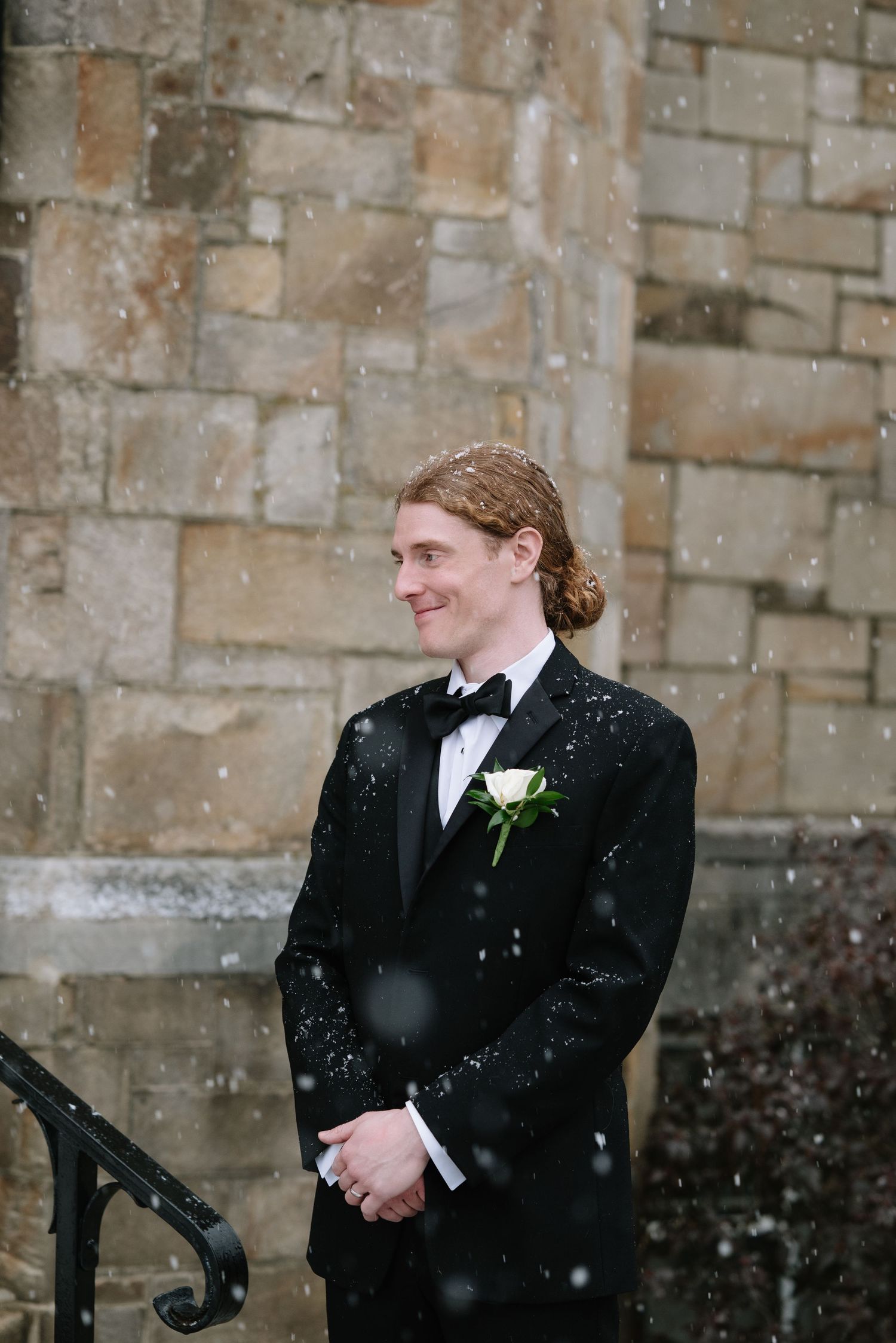 Snowy winter wedding scene with formal attire against stone church wall background.