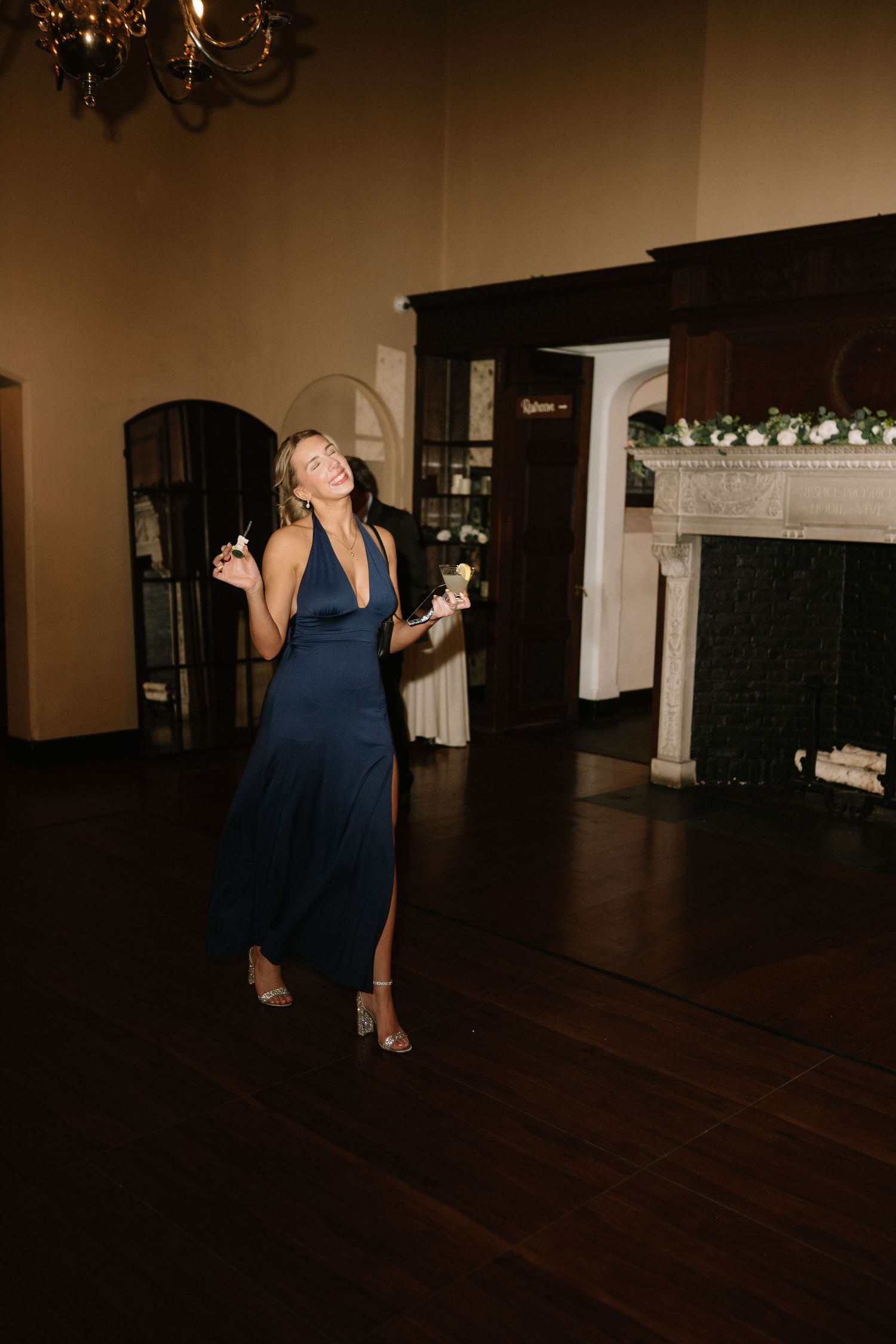 A woman in a navy blue halter dress dances joyfully in an elegant dark wood ballroom near a fireplace.