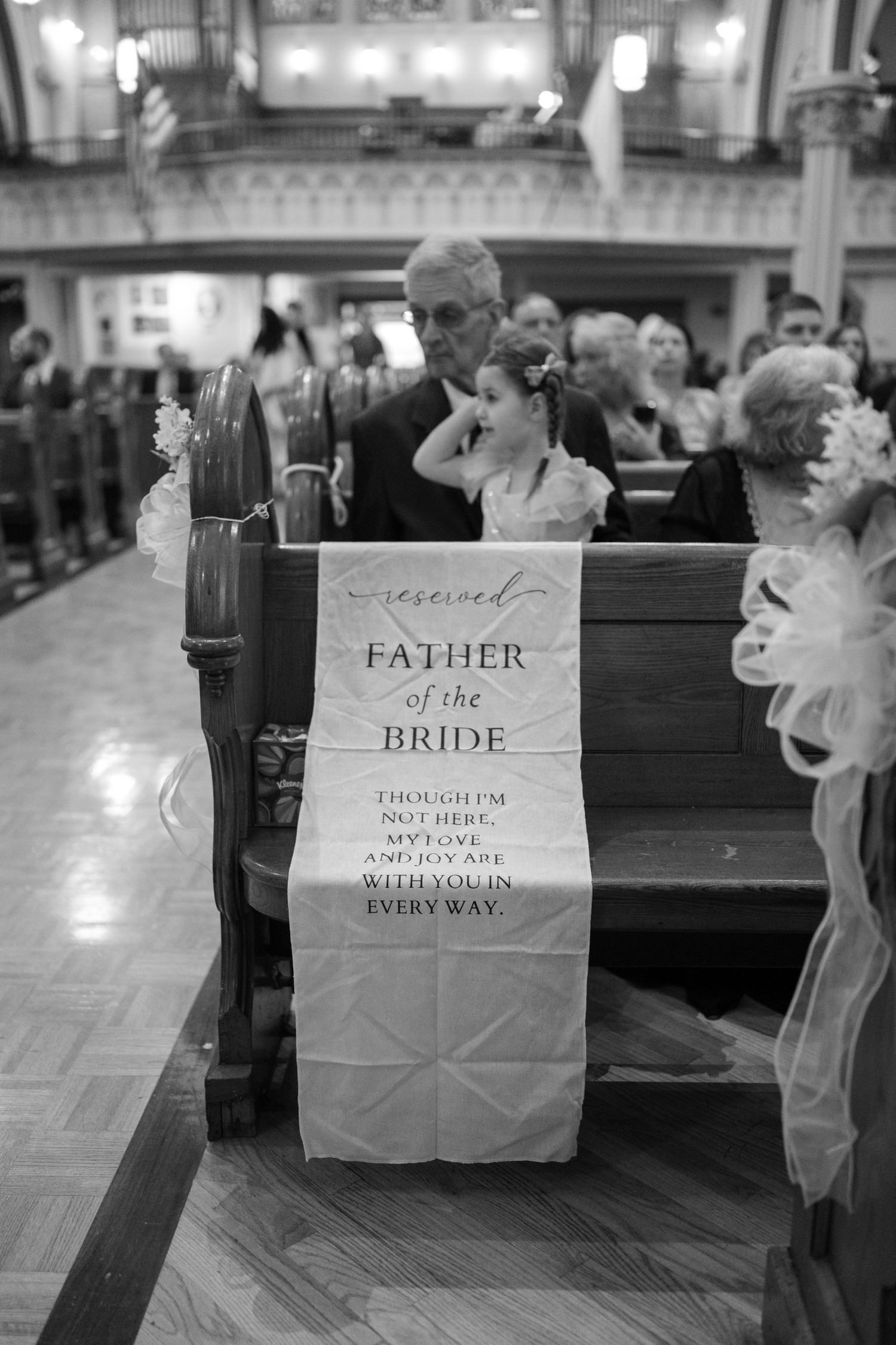 A black and white photo of a reserved church pew sign reading 'Father of the Bride' during a wedding ceremony.