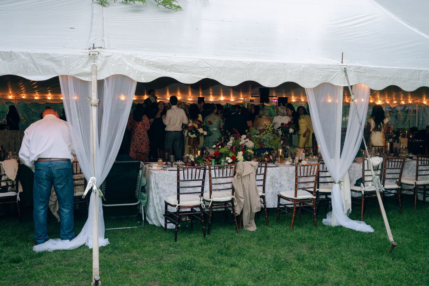 Elegant outdoor wedding reception under a white tent with string lights, draped fabric, and floral centerpieces on decorated tables.
