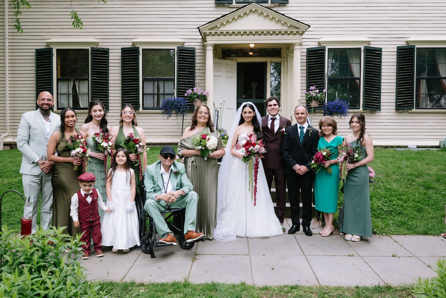 Wedding party poses in front of a white colonial house, bride in white gown with bridesmaids in green dresses.