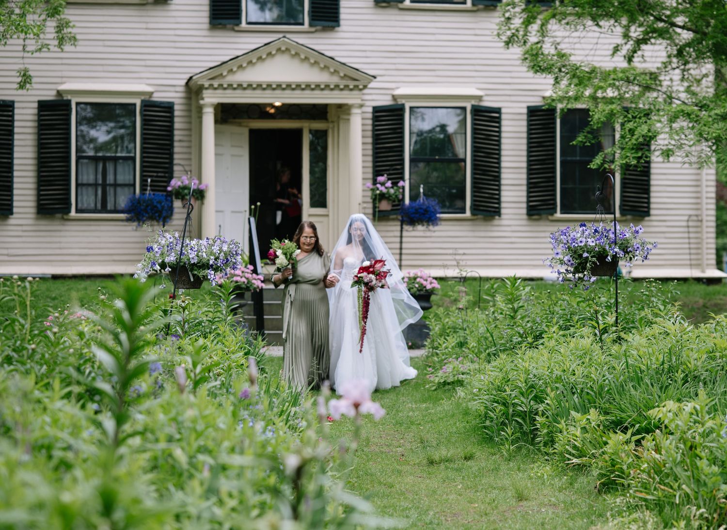 Bride in white gown walks with bridesmaid through lush garden path beside elegant colonial white house.