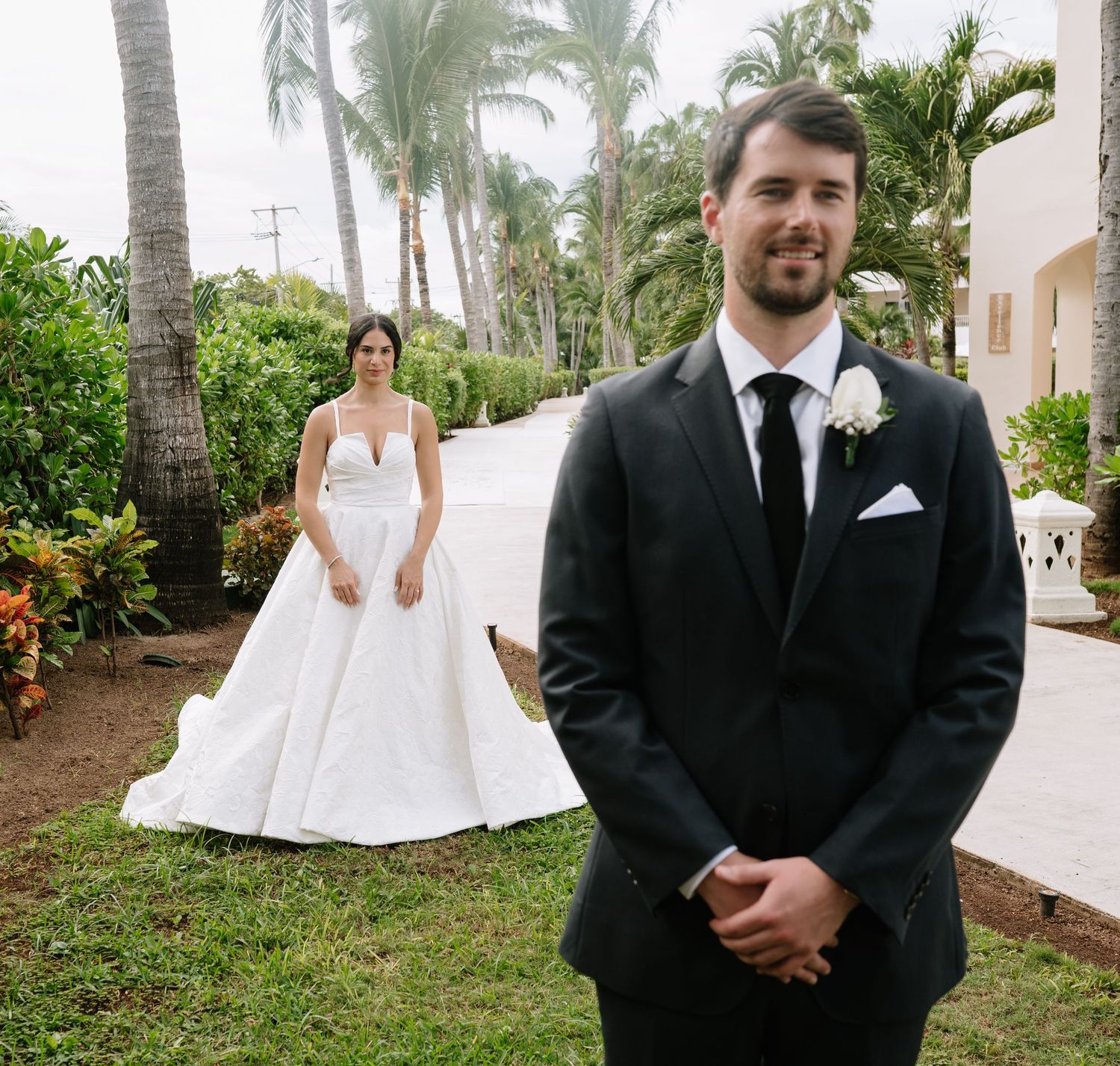 Wedding party members in formal attire pose along a palm tree-lined path at a tropical venue.