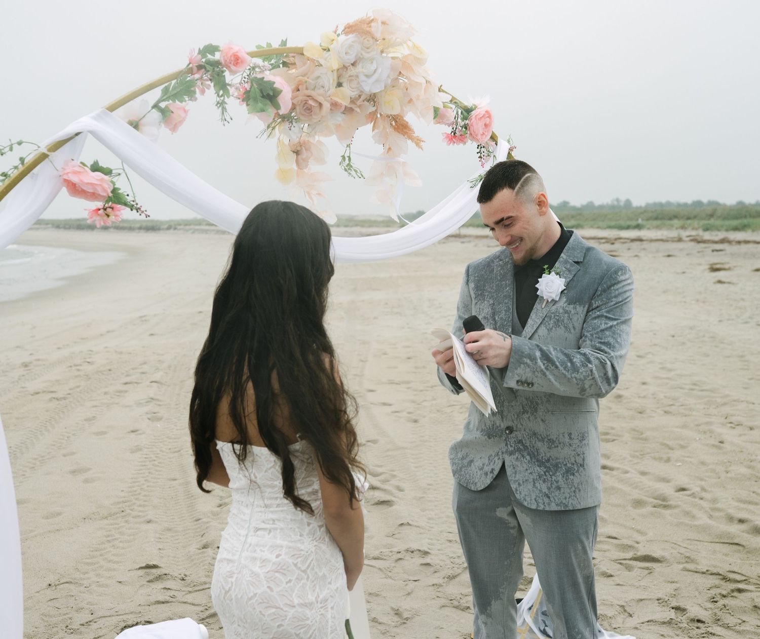 A couple shares a romantic moment under a floral arch on a cloudy beach during their wedding ceremony.