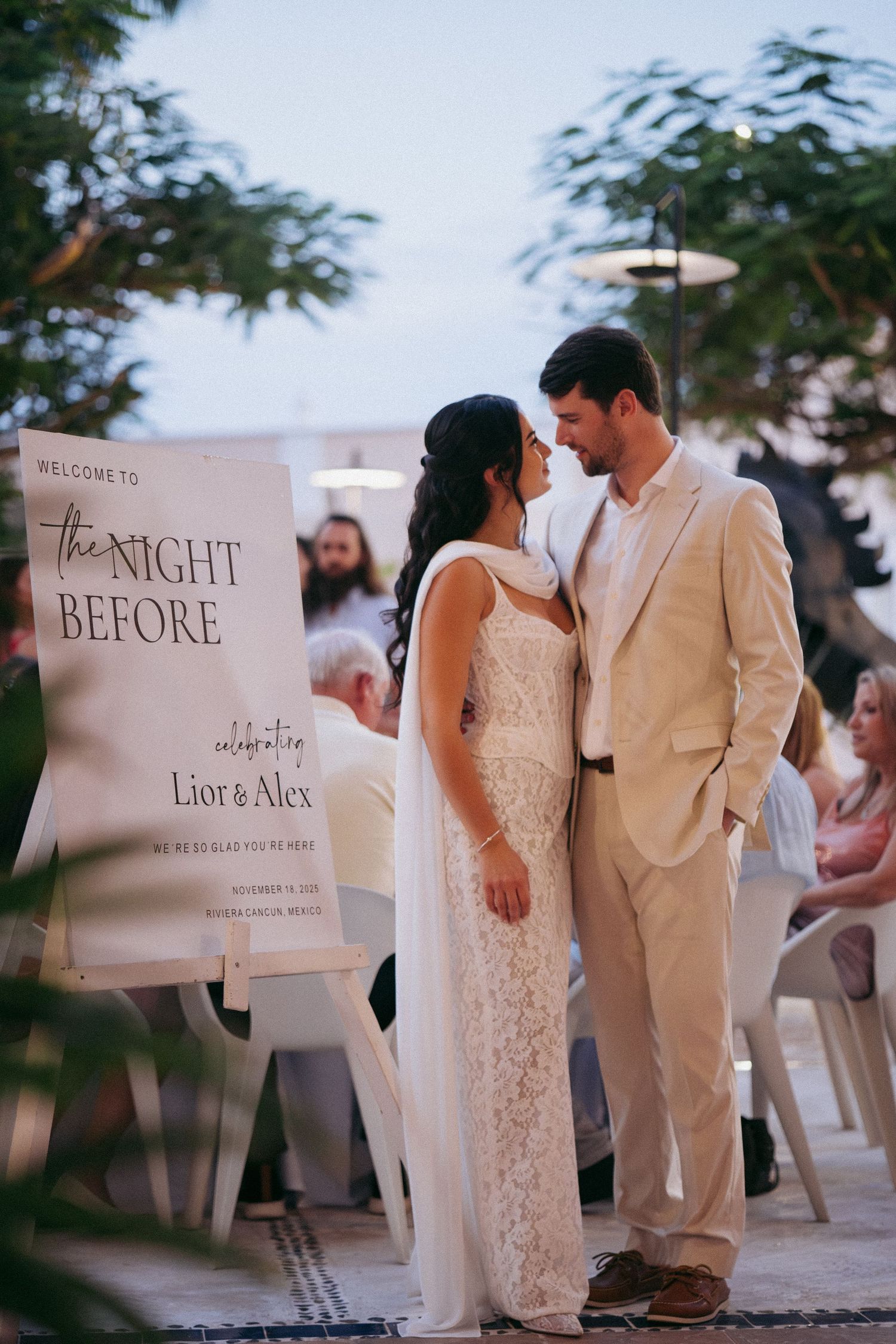 A couple in formal attire stands beside a welcome sign reading 'The Night Before' at their wedding celebration.