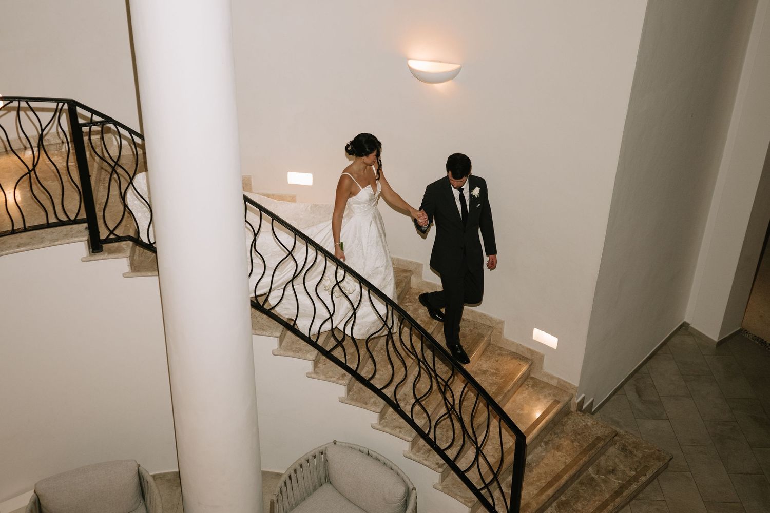 A couple in wedding attire descends an elegant curved staircase with decorative metal railings in a modern interior space.