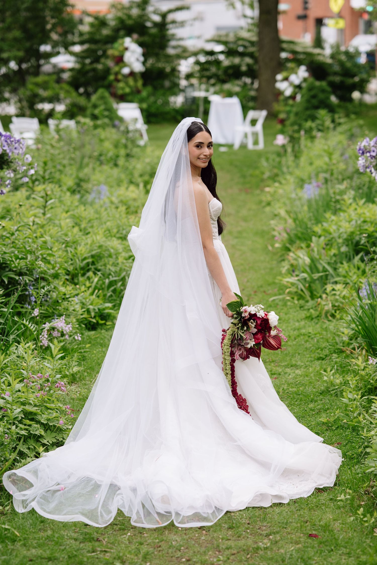 Bride in white gown and veil holding red bouquet stands in lush garden with floral arrangements in background.