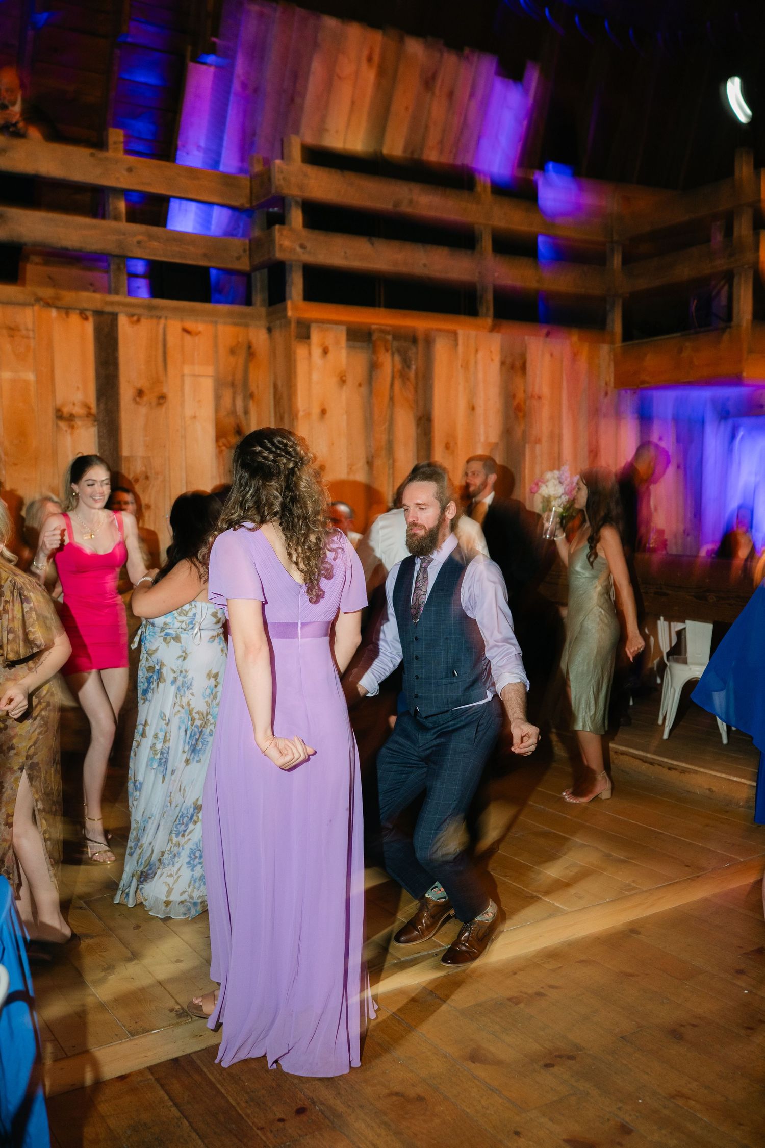 Wedding guests dancing in a rustic barn venue illuminated by colorful purple lighting.