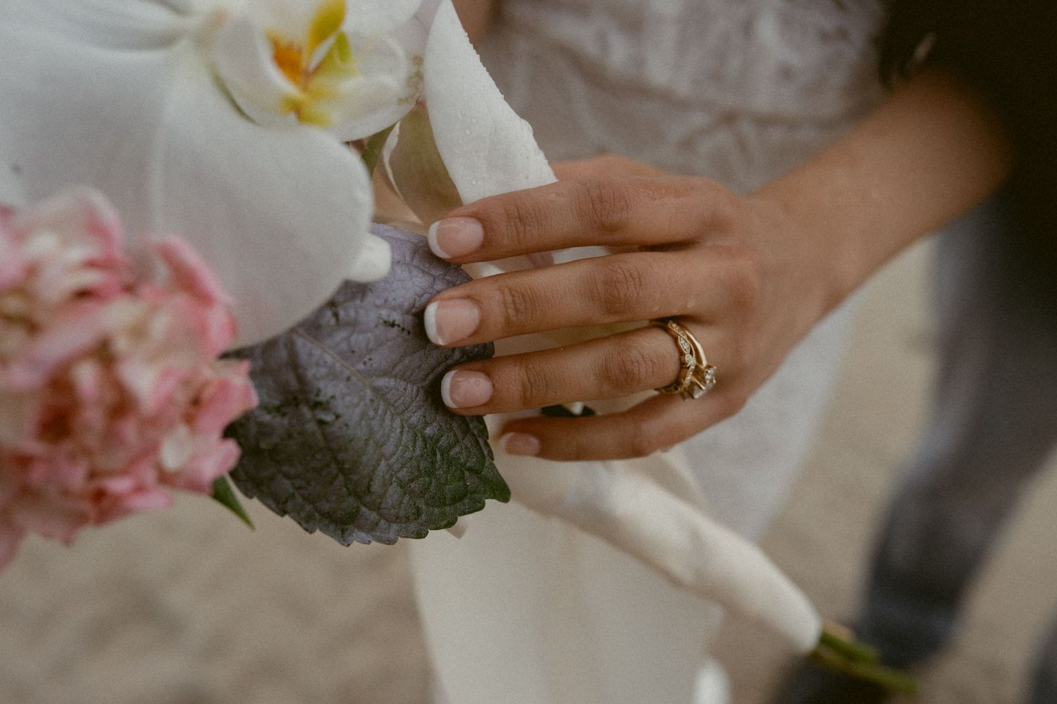 Close-up detail of hands holding delicate white and pink wedding flowers with gold rings.