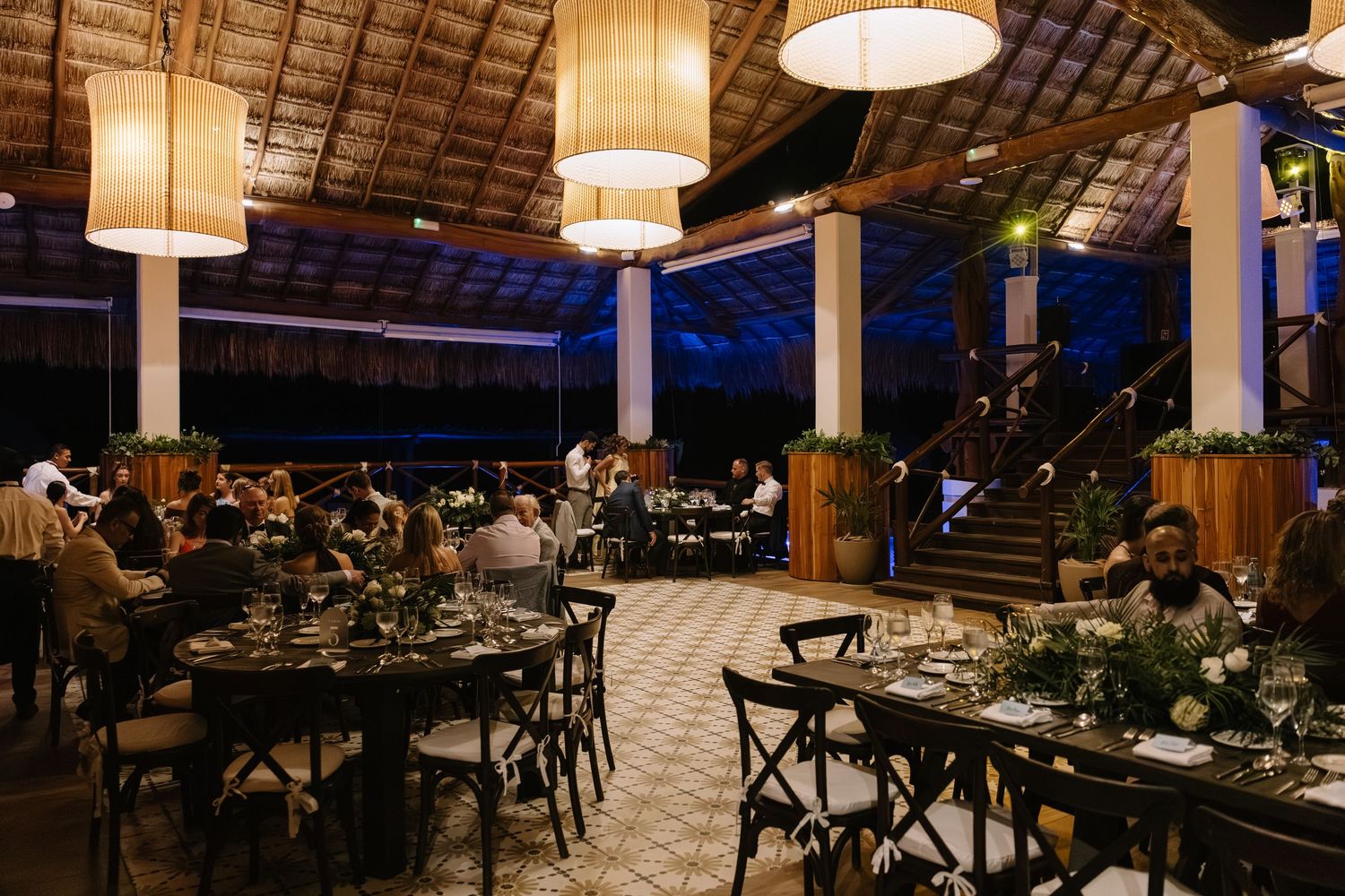 Elegant wedding reception venue with round tables, cross-back chairs, and large pendant lights under a vaulted ceiling.