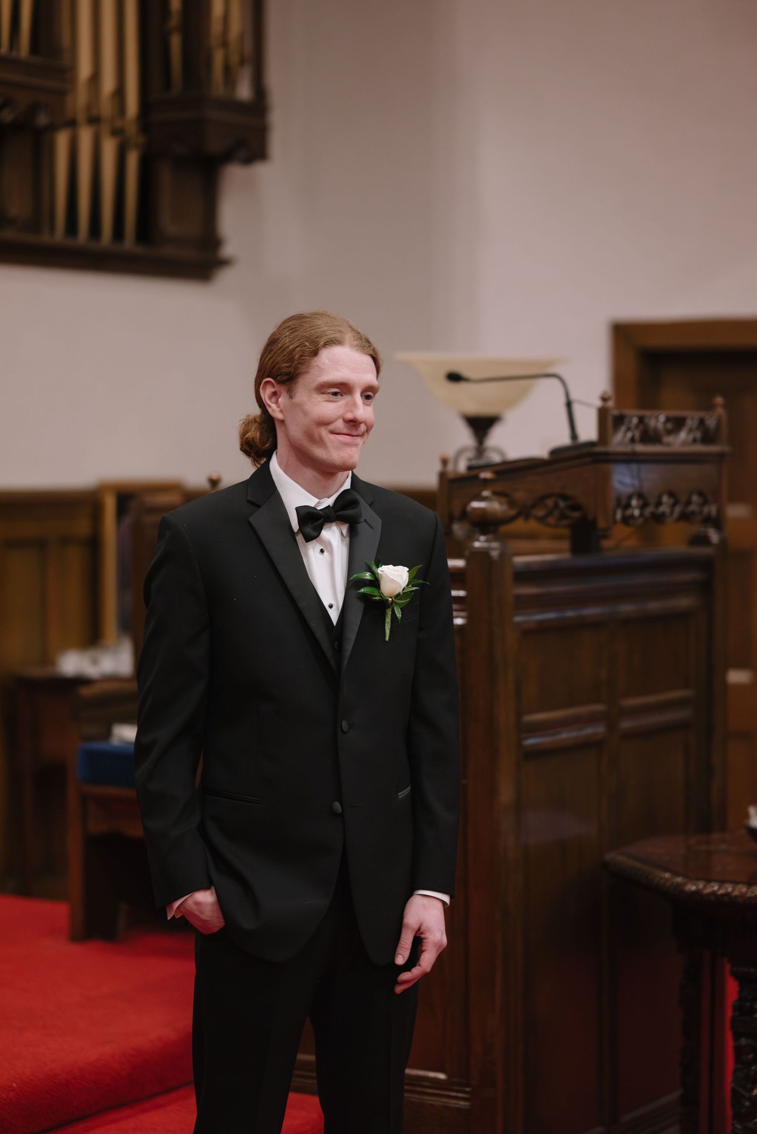 A person in a black tuxedo and bow tie stands in a church sanctuary with red carpeting and wooden pews.