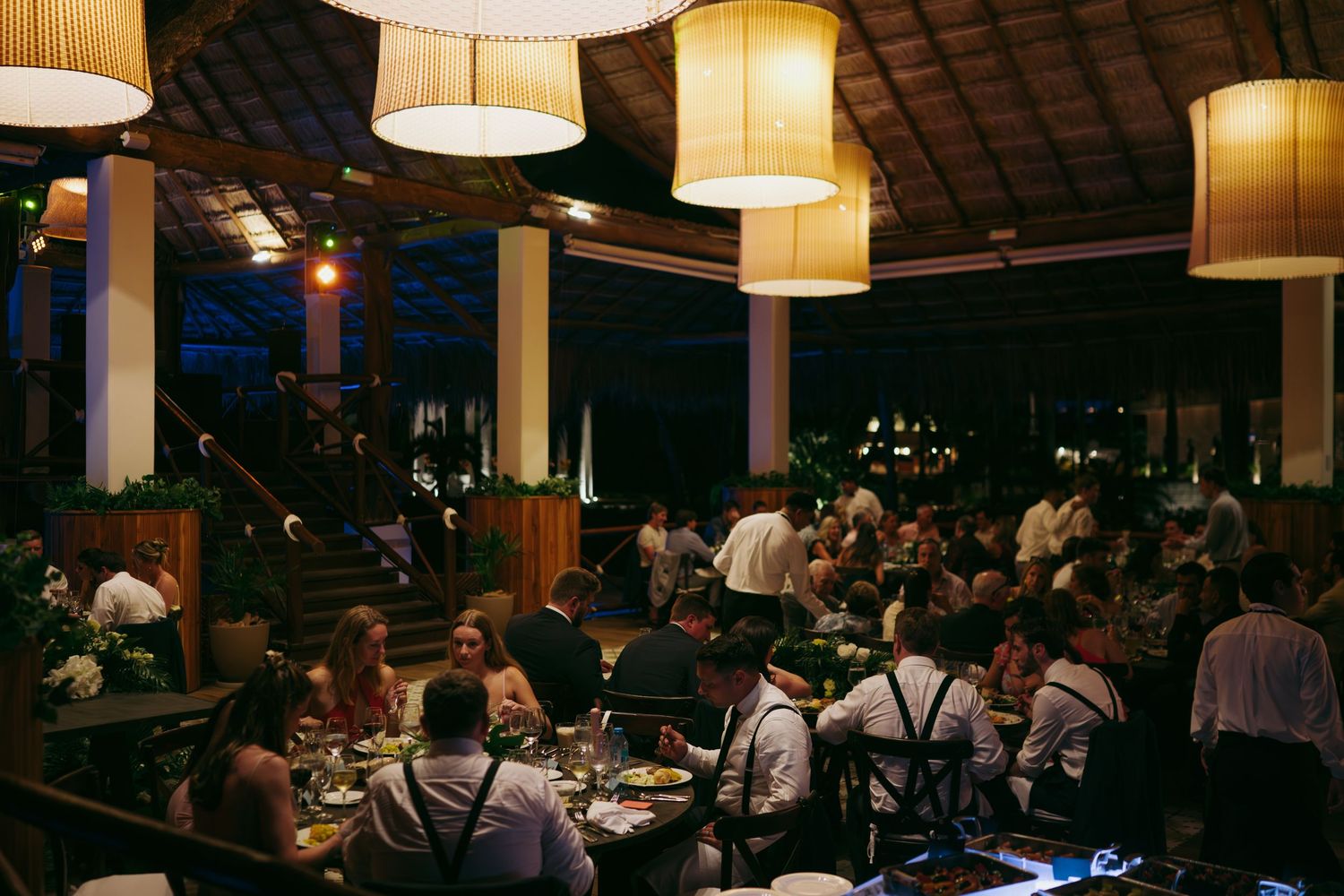 Elegant reception venue with warm lighting from drum pendant lamps and guests dining at round tables.