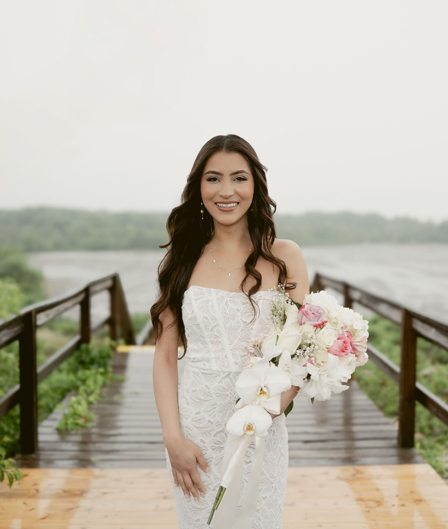 A beautiful white wedding dress and pink floral bouquet photographed on a wooden boardwalk by the water.