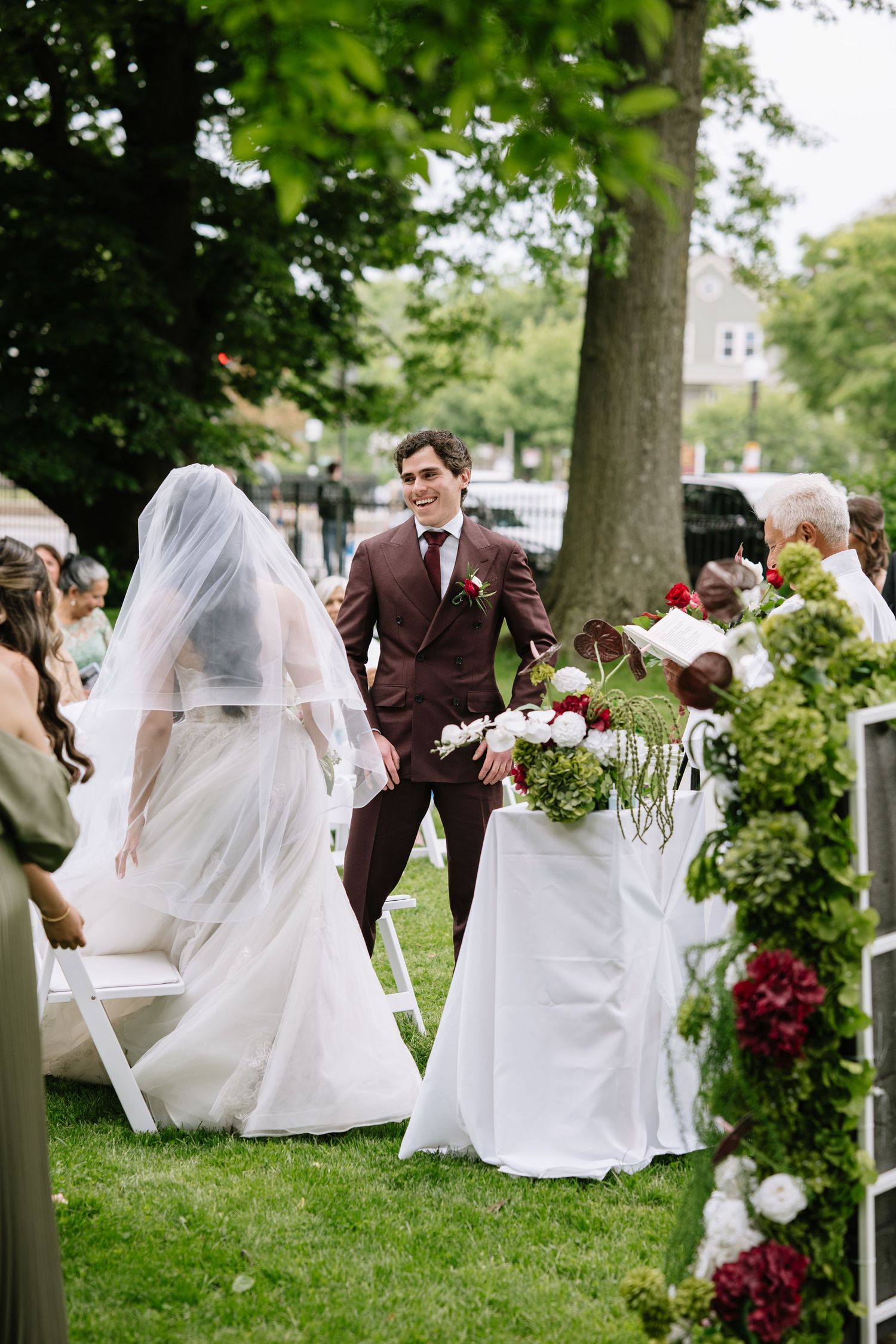 Groom in burgundy suit smiles as bride walks down outdoor wedding aisle lined with floral arrangements.