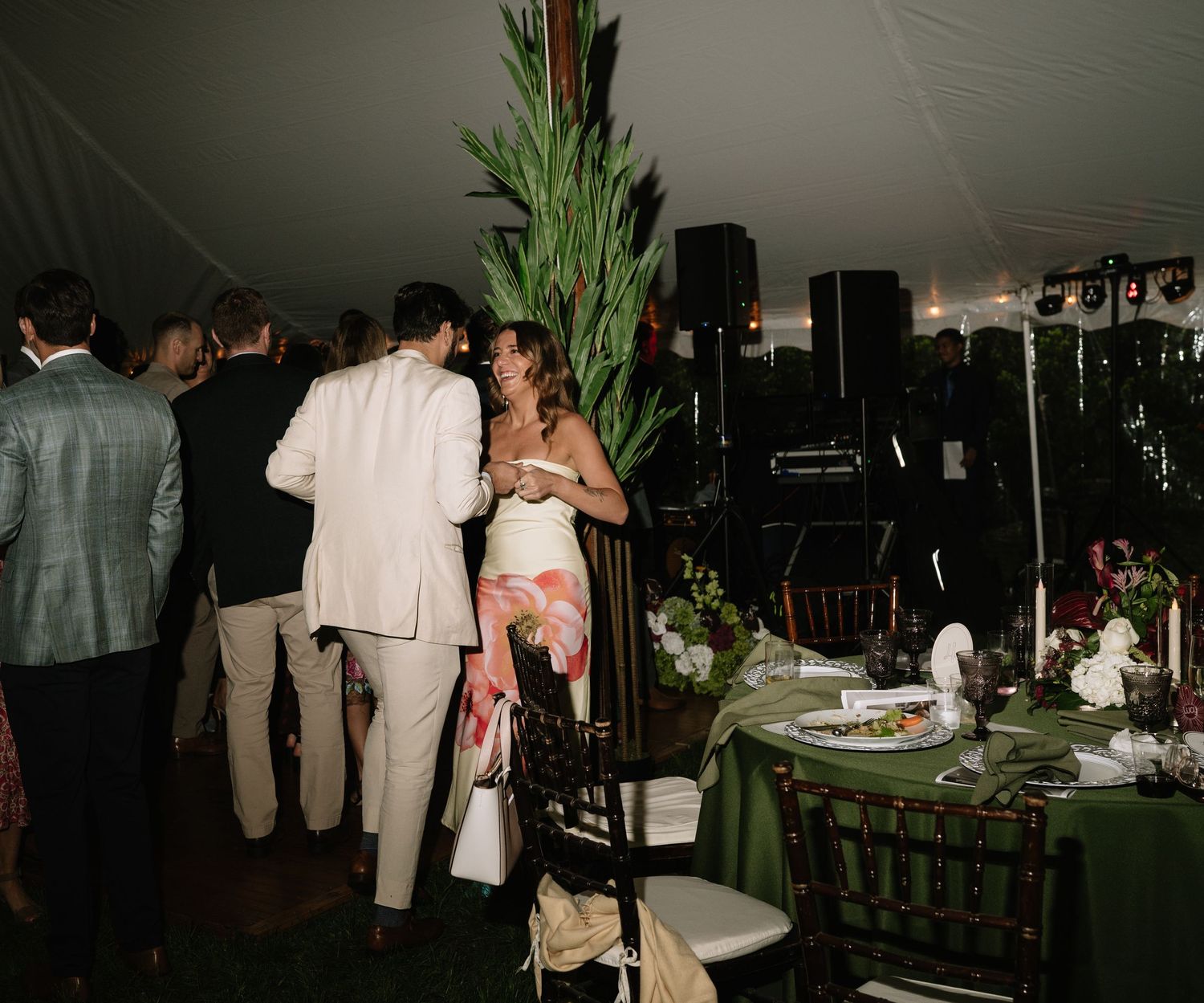 Guests mingle under a white tent at an elegant outdoor wedding reception with green tablecloths and tropical floral arrangements.