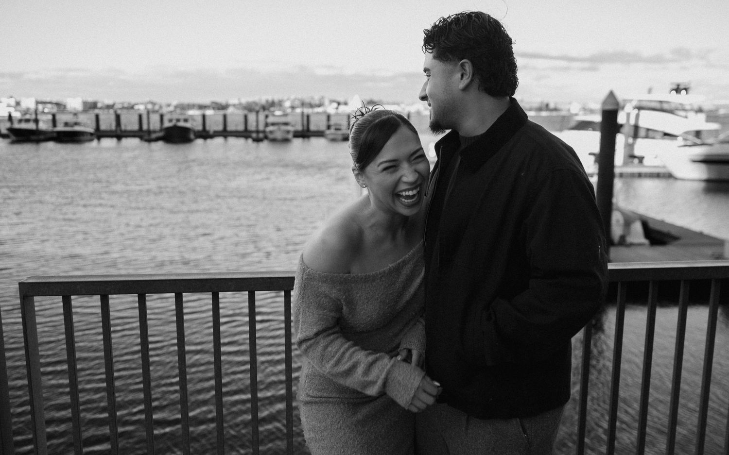 Black and white photo of a couple sharing an intimate moment by a waterfront dock.