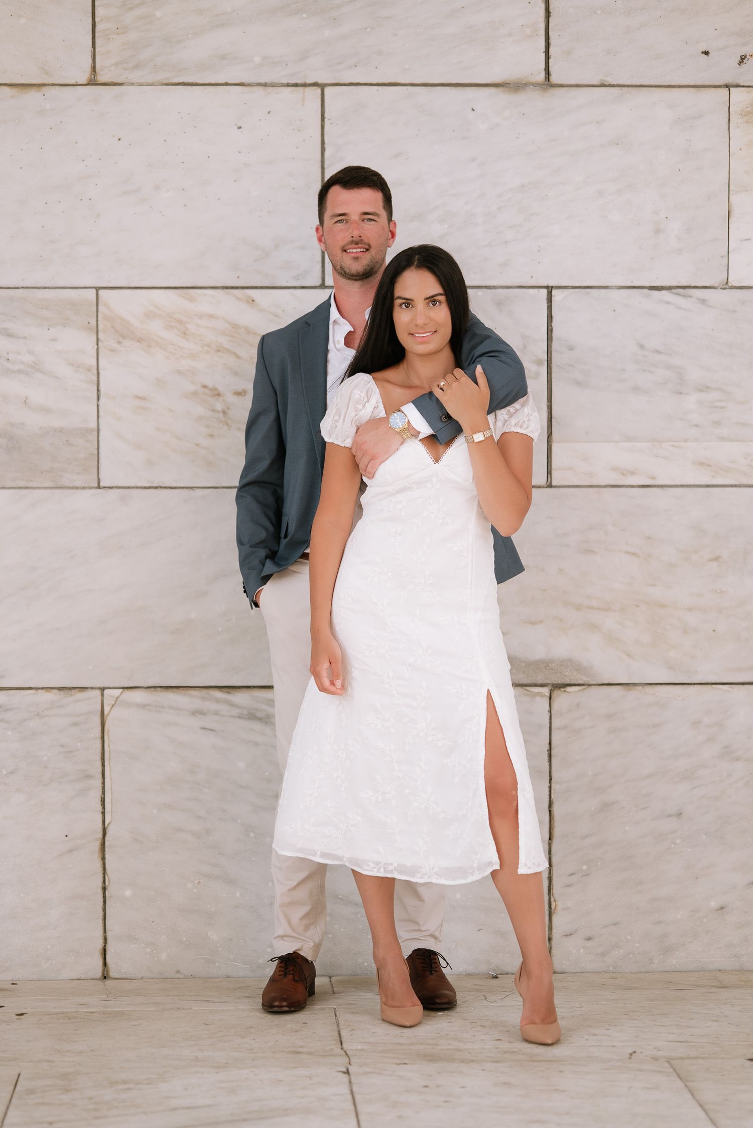 Romantic engagement photo shoot with couple in white dress and grey blazer against stone wall backdrop.