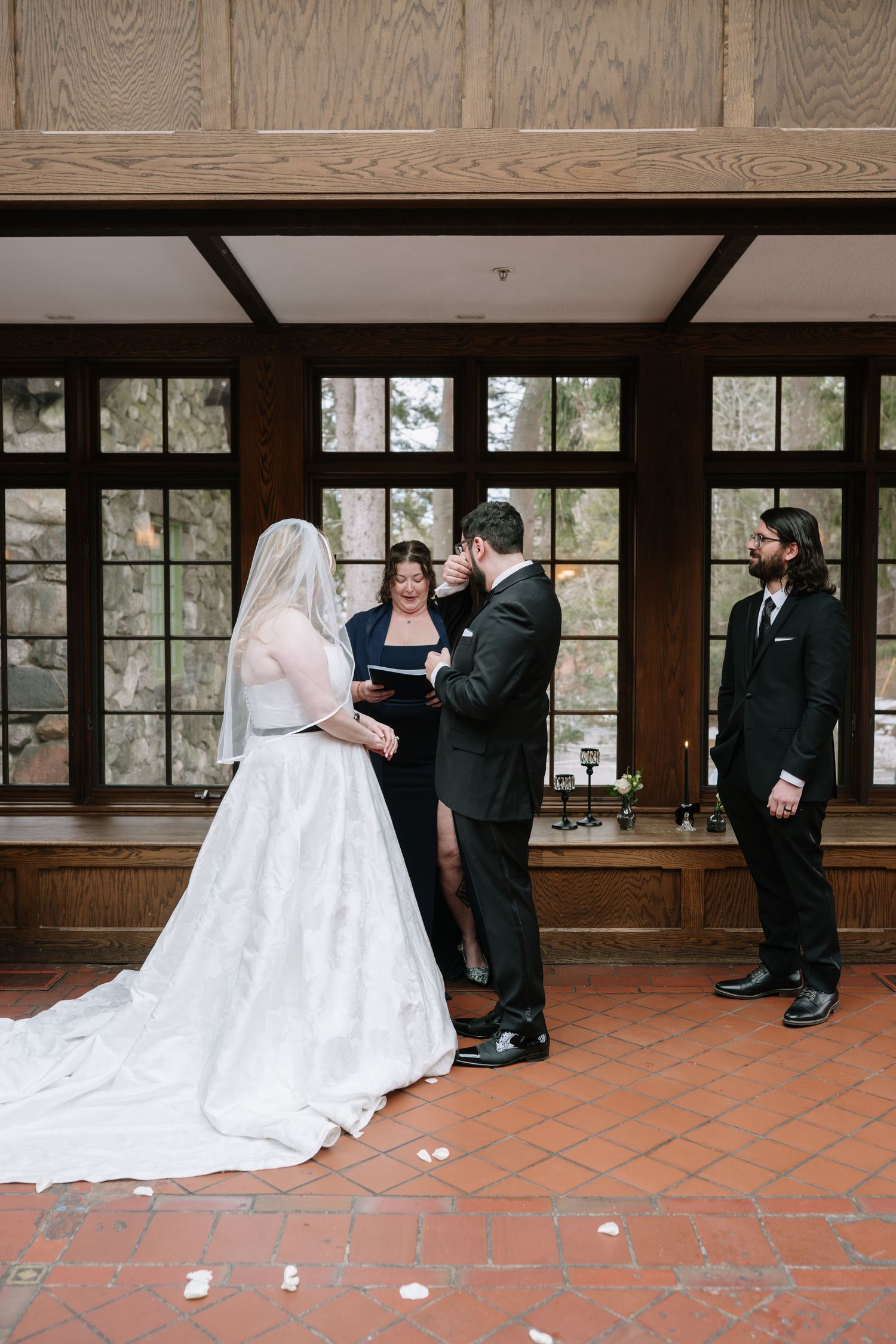 Bride and groom exchange vows with officiant and groomsman in elegant wood-paneled venue with large windows.