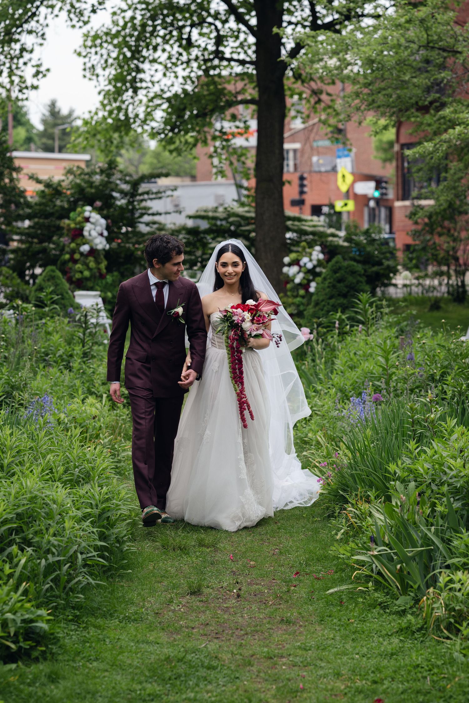Bride and groom walk through lush garden path, she in white gown with bouquet, he in burgundy suit.