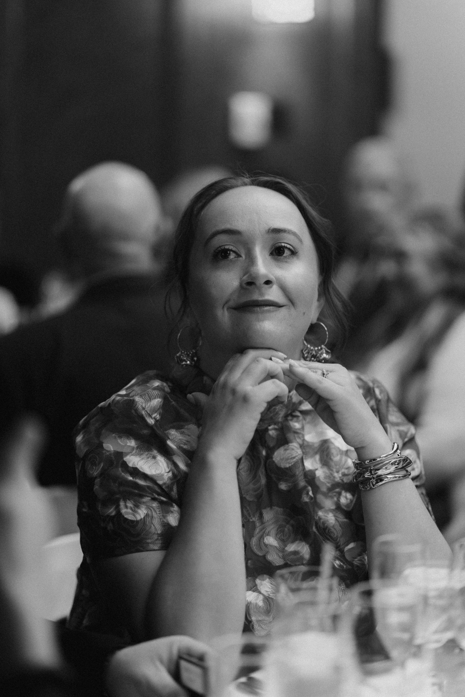 Black and white photo of a woman with hoop earrings and bracelets sitting at a dinner table, hands clasped, looking up.